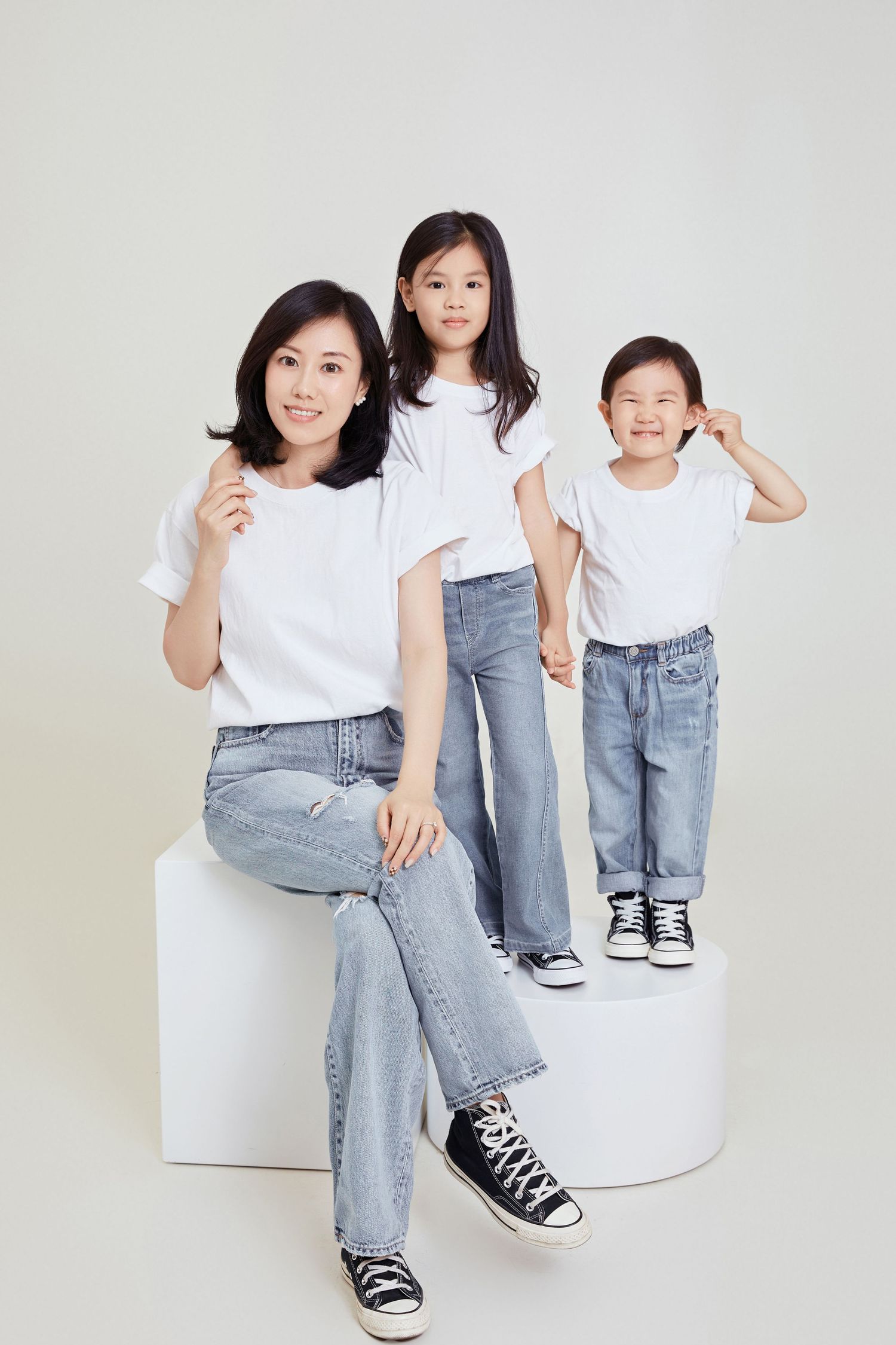 Asian mother and two daughters in matching white t-shirts and jeans pose together in a bright studio.