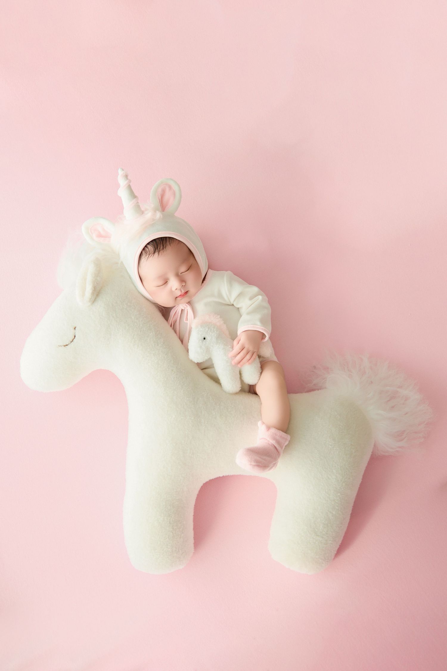 Newborn baby sleeping on a white unicorn stuffed animal toy, wearing a unicorn hat on a pink background.