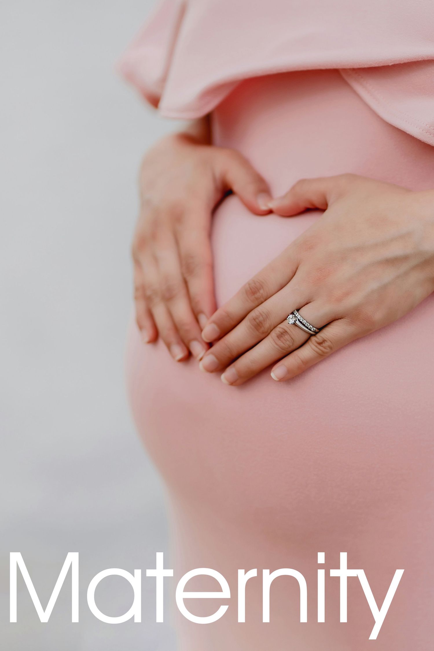 Pregnant woman in pink dress forming heart shape with hands on baby bump, wearing wedding ring, maternity photo.