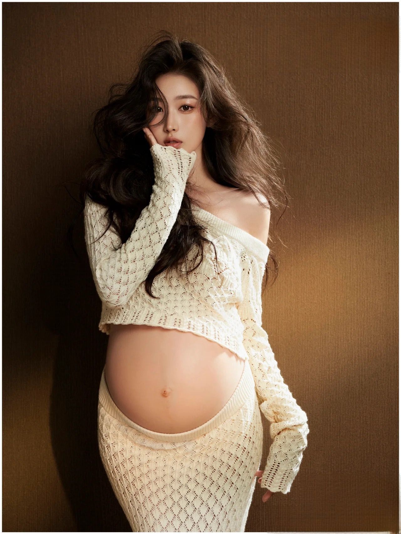 Elegant pregnant woman posing in cream off-shoulder knit two-piece outfit for a stylish maternity shoot.