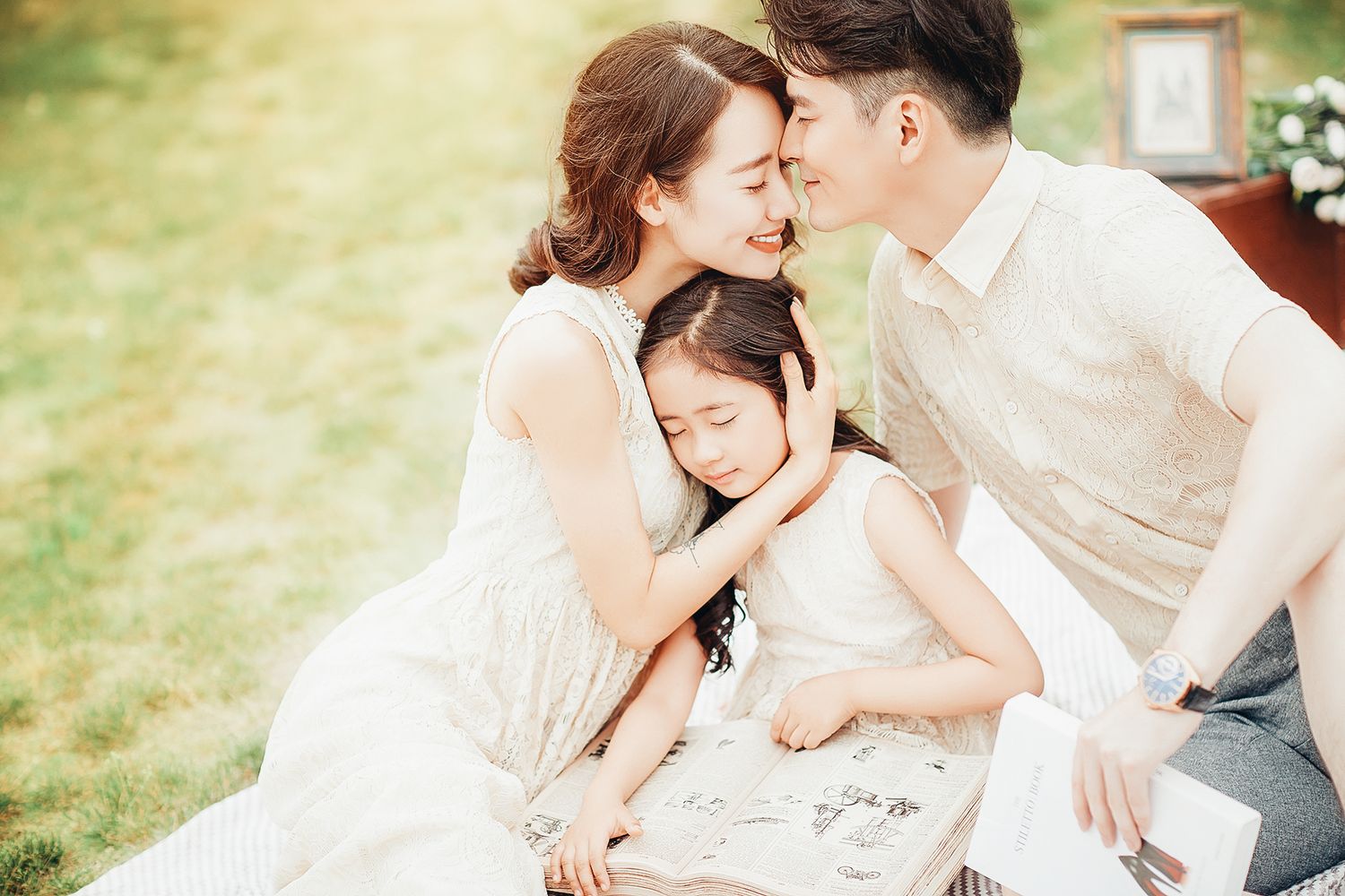 A loving Asian family shares a tender moment as parents kiss their young daughter on a green lawn.