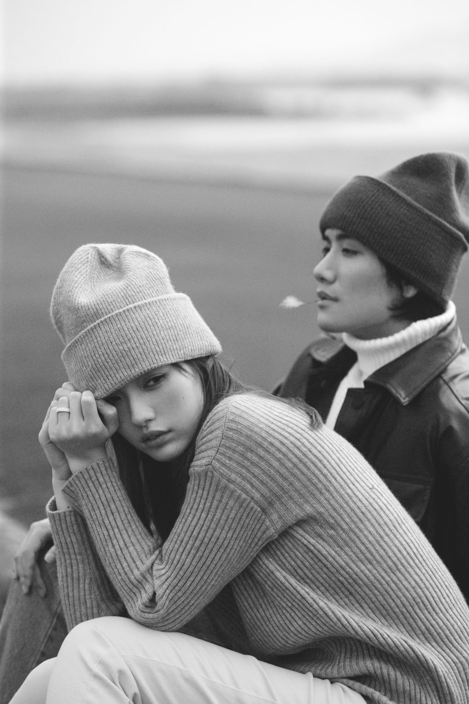 Black and white photo of a young couple in knit beanies and sweaters sitting together near a waterfront.