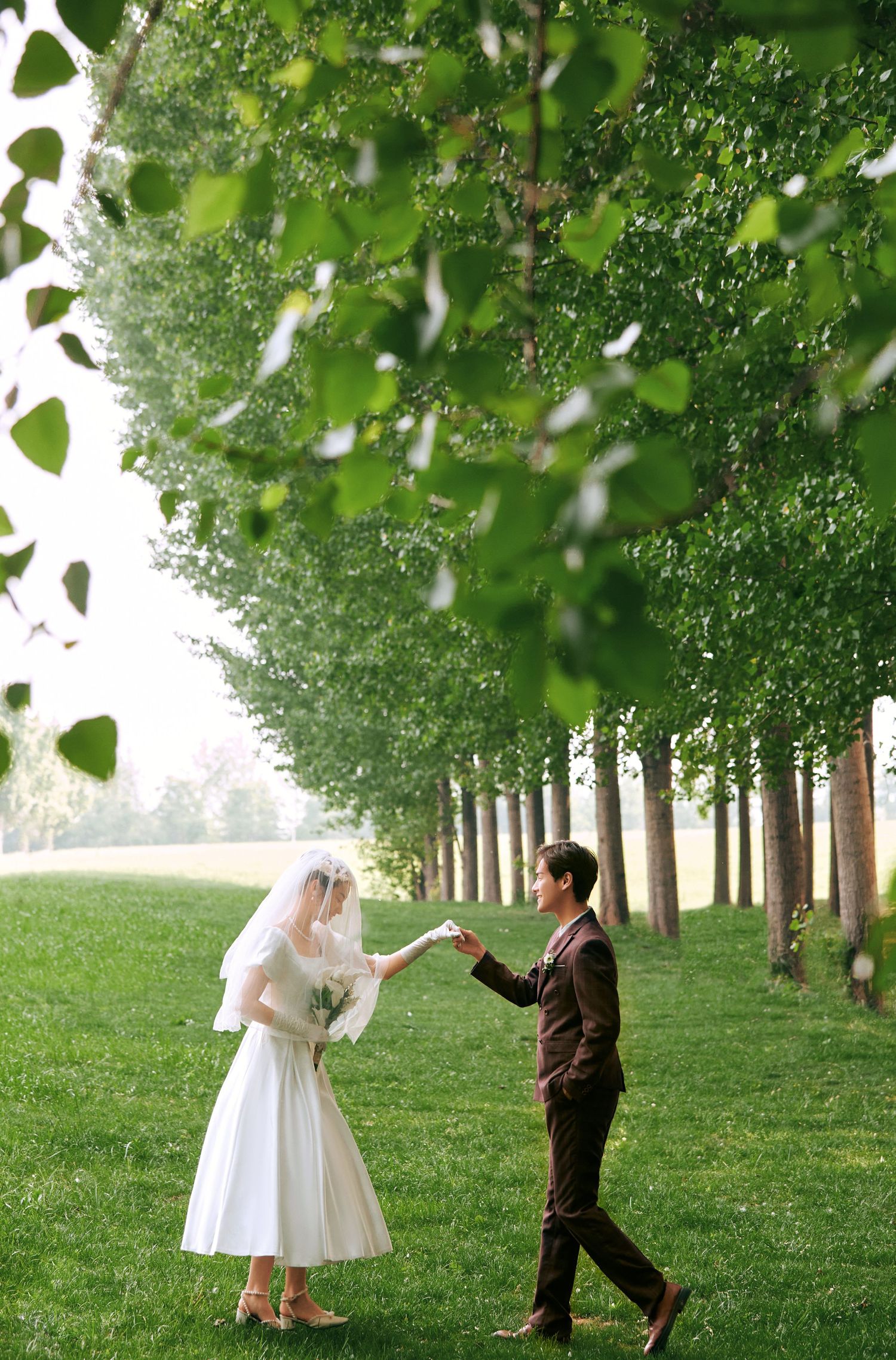 Bride and groom holding hands in lush green tree-lined park during outdoor wedding photo session.