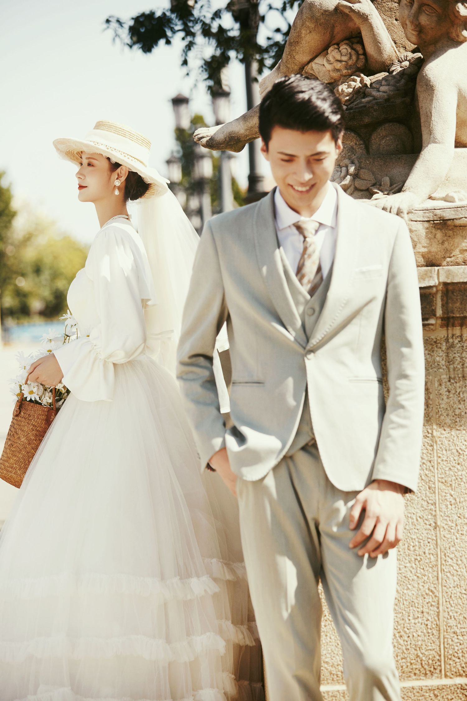 Elegant Asian couple in vintage-style wedding attire pose near stone sculpture in sunny outdoor setting.