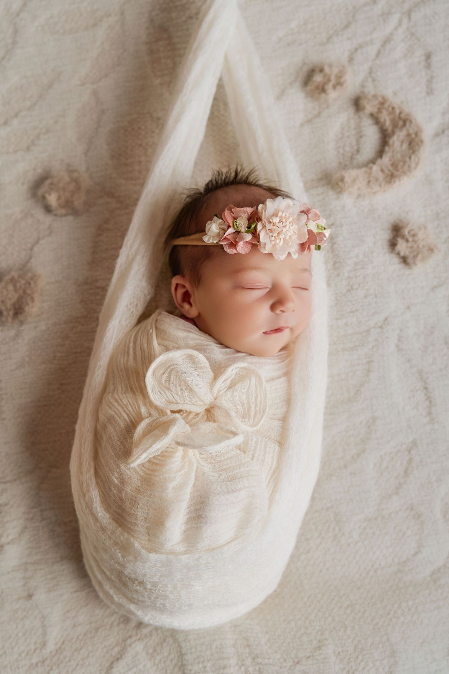 Newborn baby girl swaddled in white wrap with pink floral headband, sleeping peacefully on soft cream blanket.