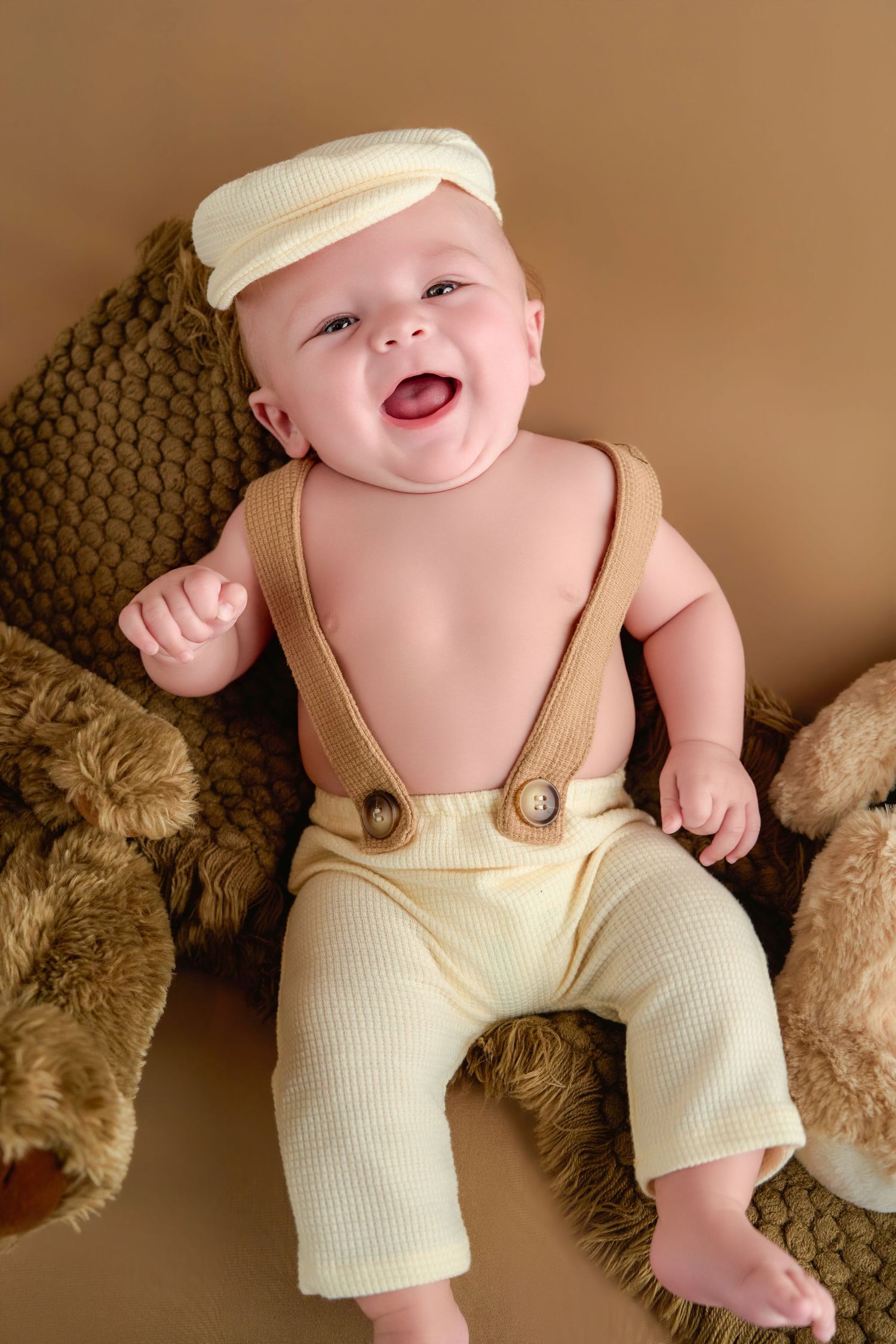 Smiling baby wearing cream suspender pants and a white cap, surrounded by teddy bears on a brown knit blanket.