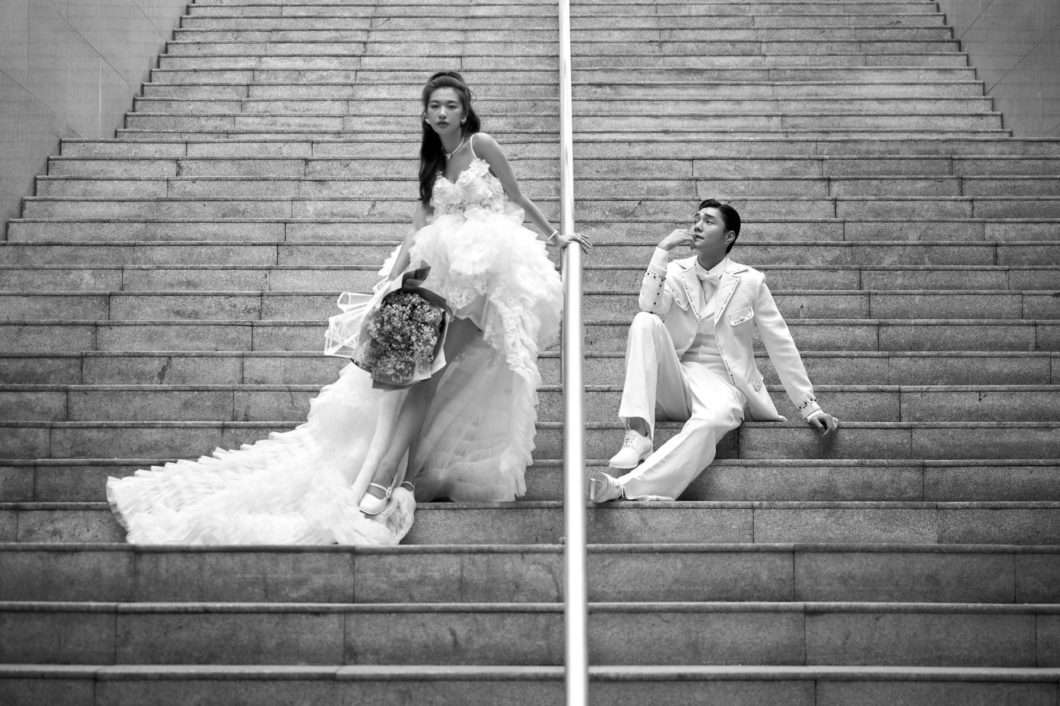 Black and white photo of bride and groom posing on grand staircase in elegant wedding attire.