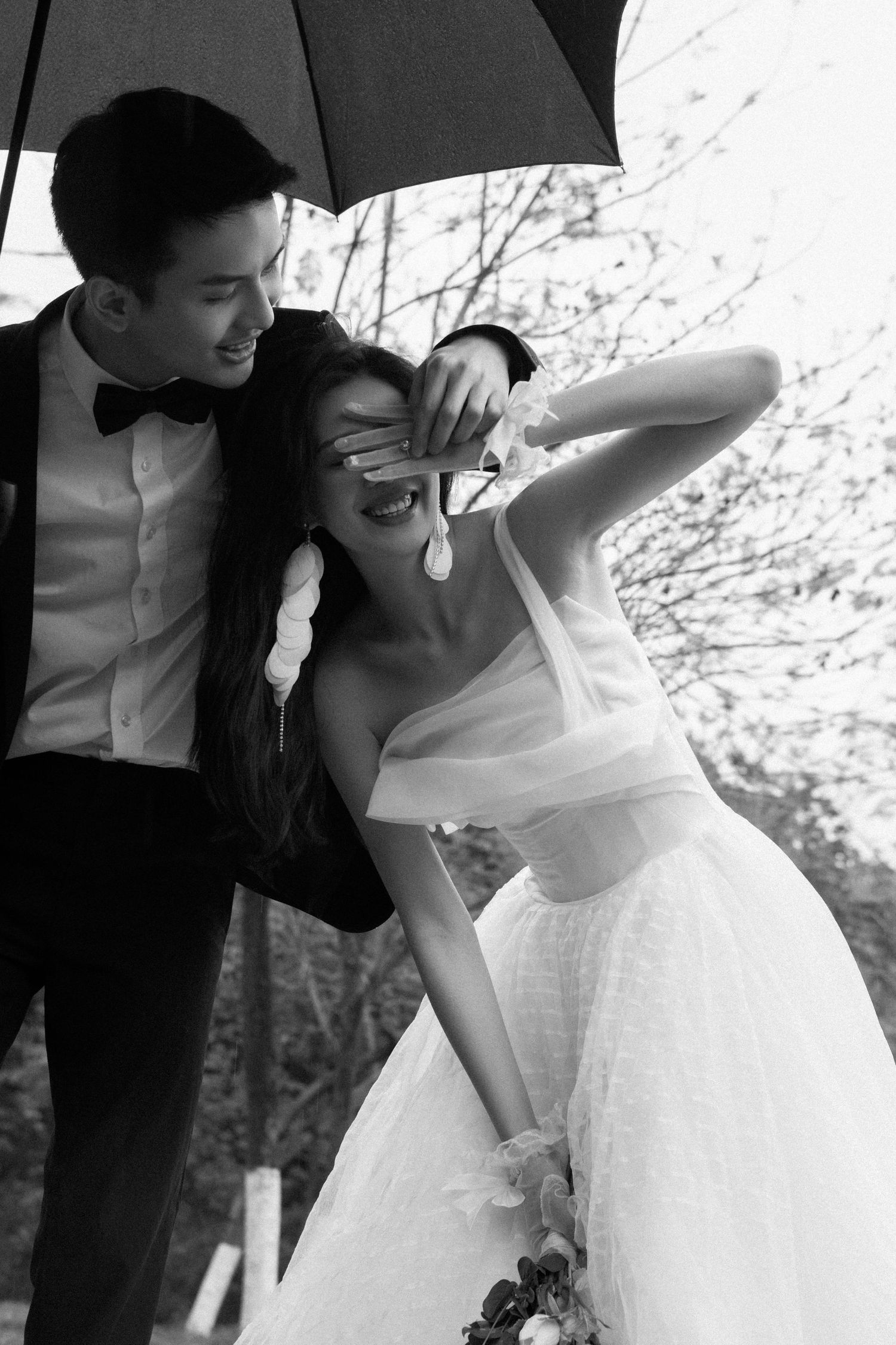 Playful black and white wedding photo of groom covering brides eyes while sharing a joyful moment under an umbrella.