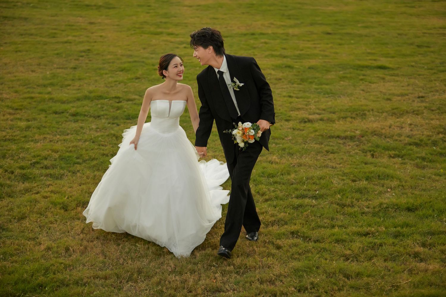 A bride in a white gown and groom in a black suit walk hand-in-hand across a green field.