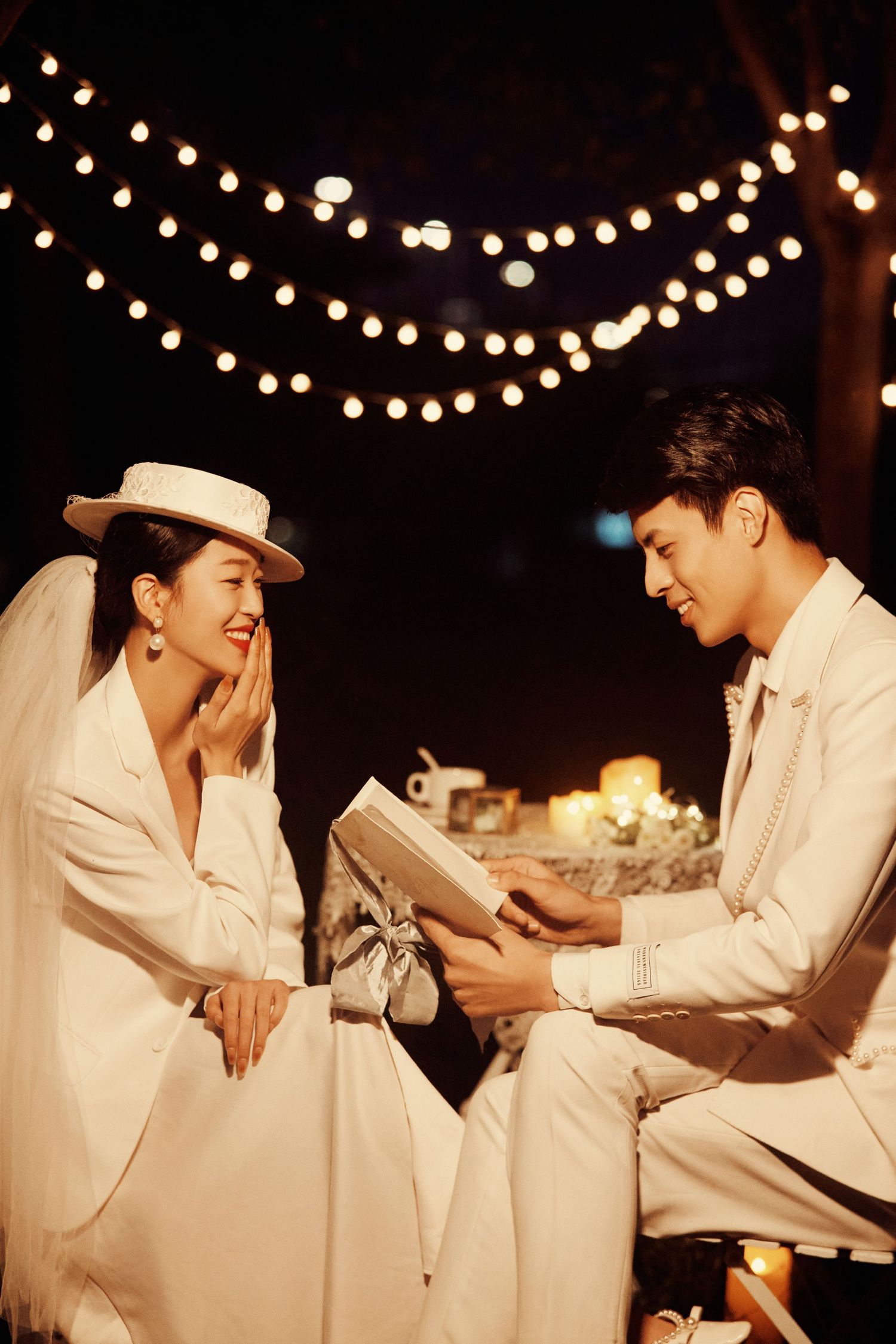 Joyful bride in white hat and groom exchanging gifts under glowing string lights in nighttime setting.
