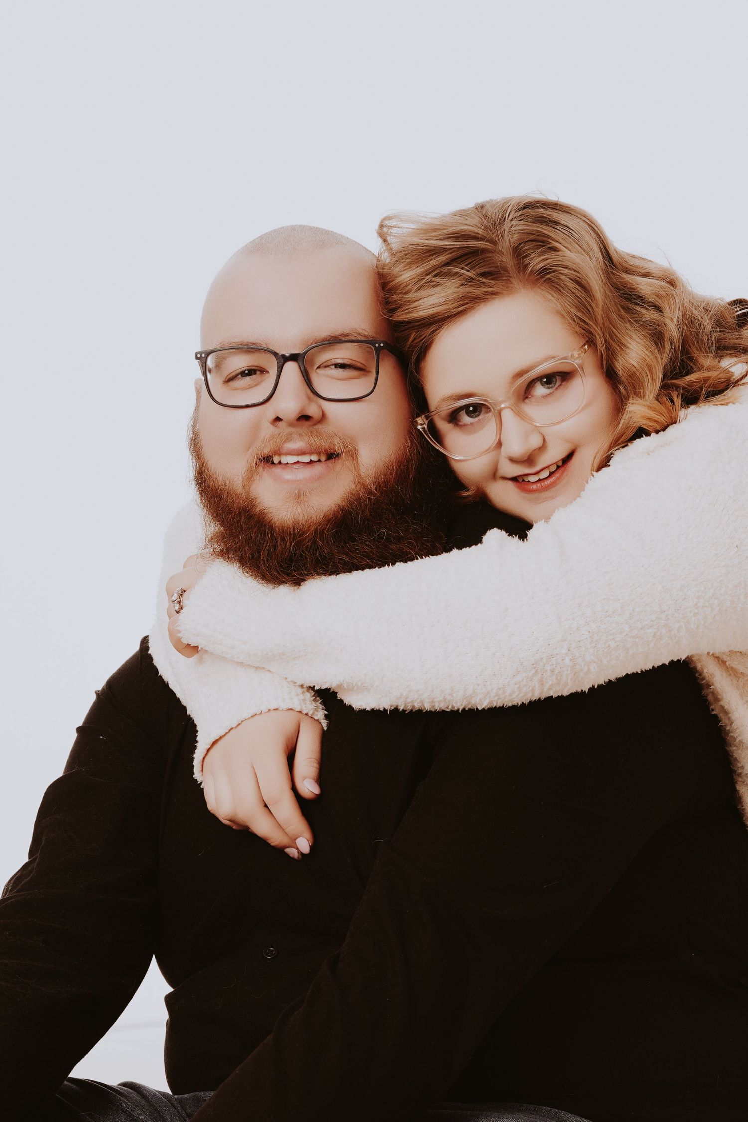Smiling bald bearded man with glasses embraced from behind by blonde curly-haired woman wearing white jacket.