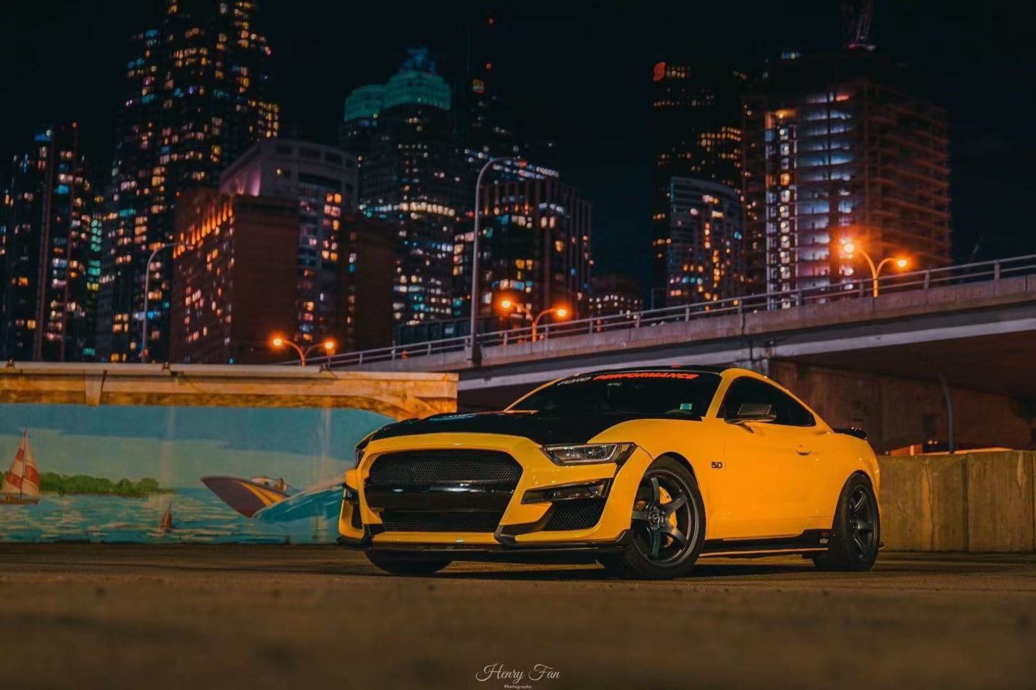 A yellow Ford Mustang GT parked at night with a glowing city skyline and bridge lights reflecting in the background.
