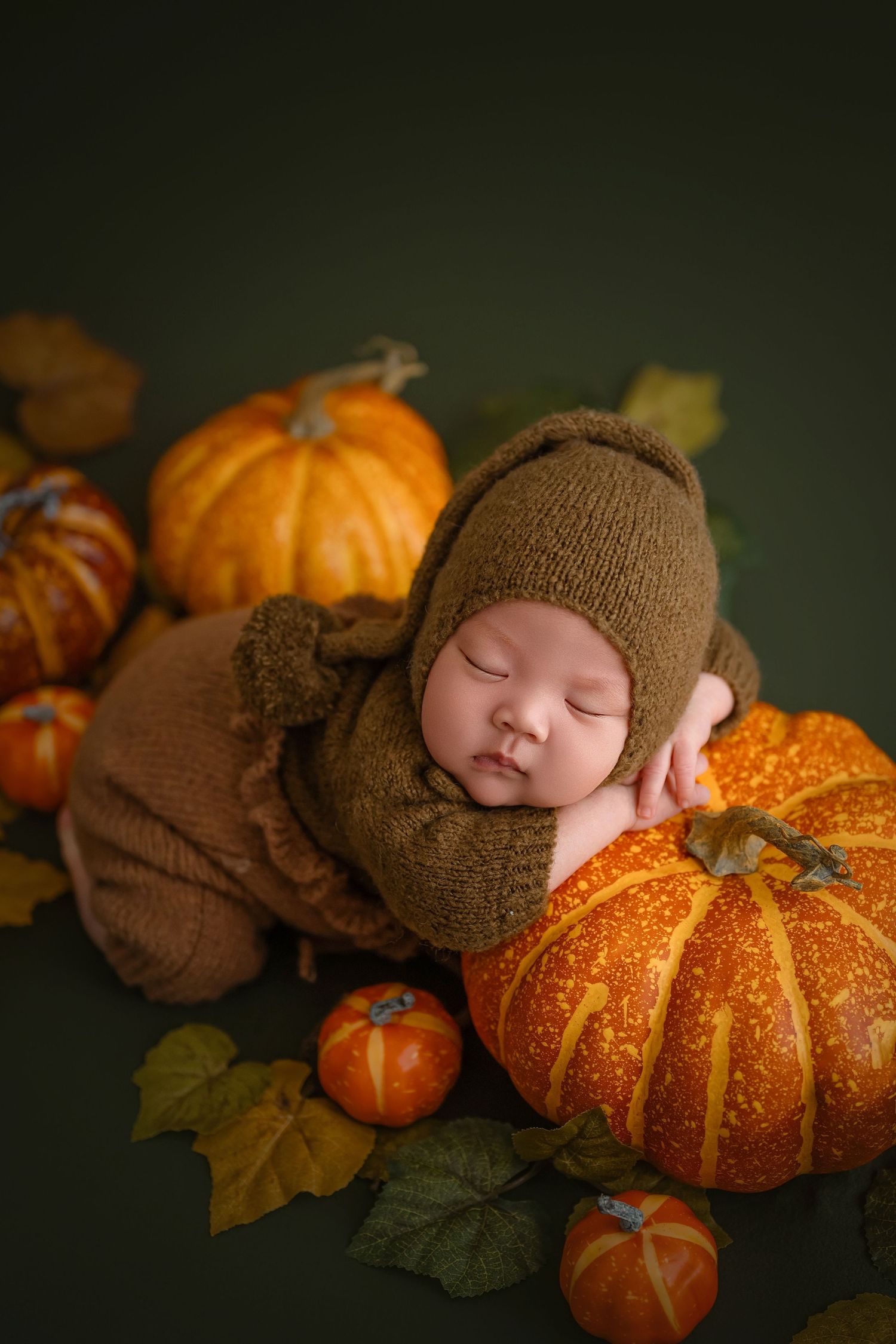 Sleeping newborn baby in brown knit outfit resting on orange pumpkin with fall leaves on green background.