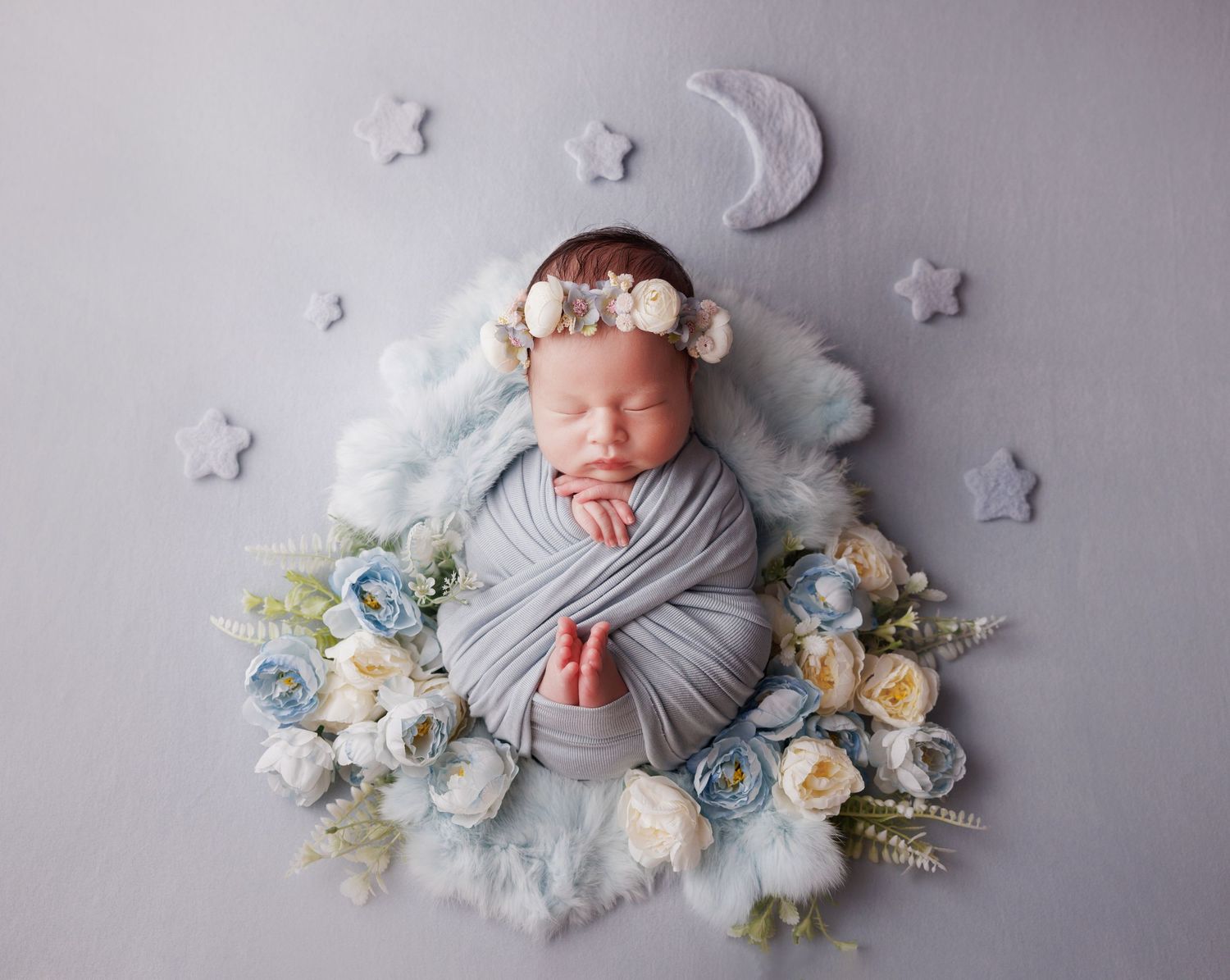 Swaddled newborn baby with floral crown sleeping in blue fluffy nest with roses, stars, and moon decorations.