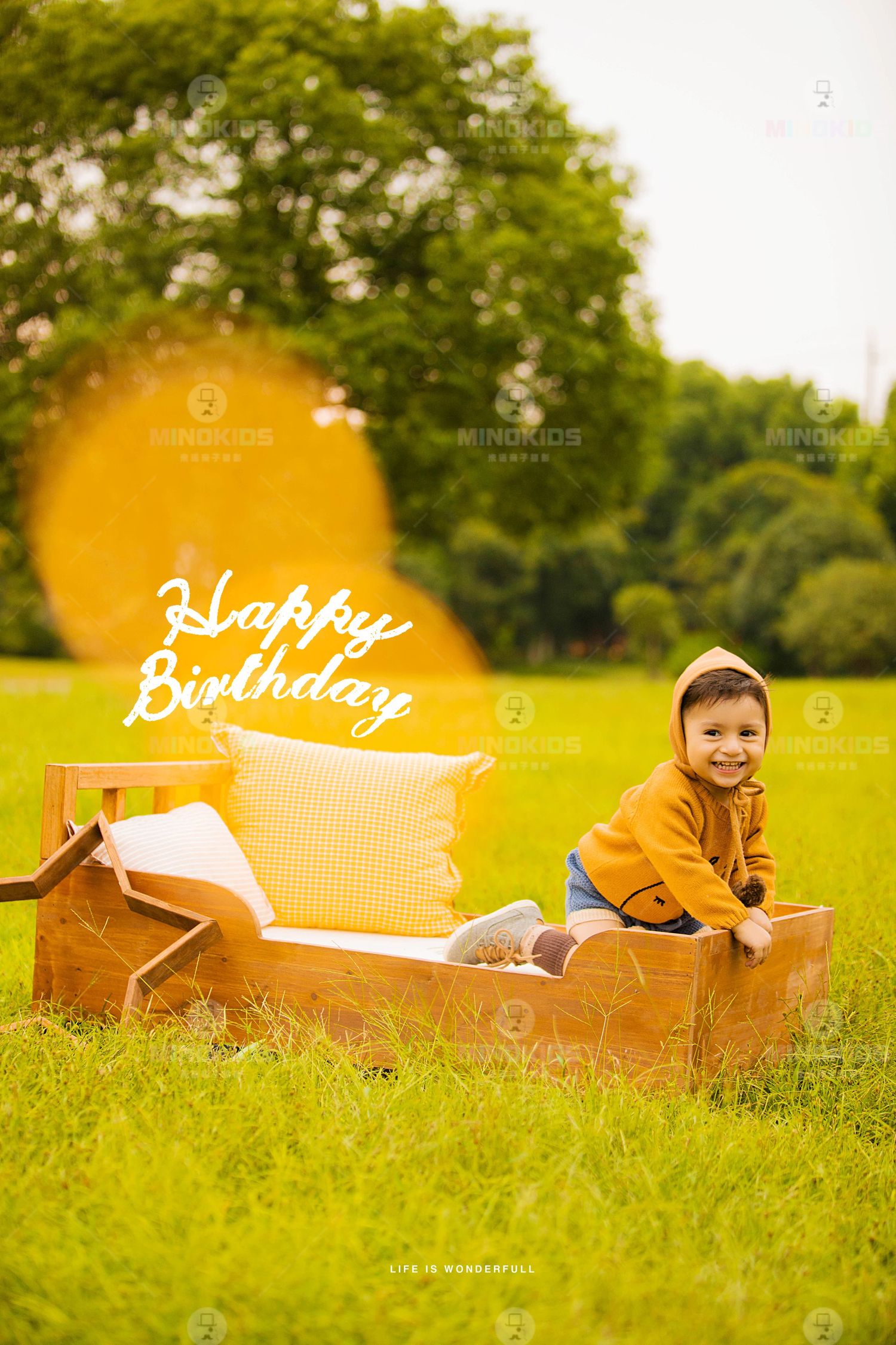 Smiling toddler in yellow jacket poses beside wooden bed on green grass with Happy Birthday text.