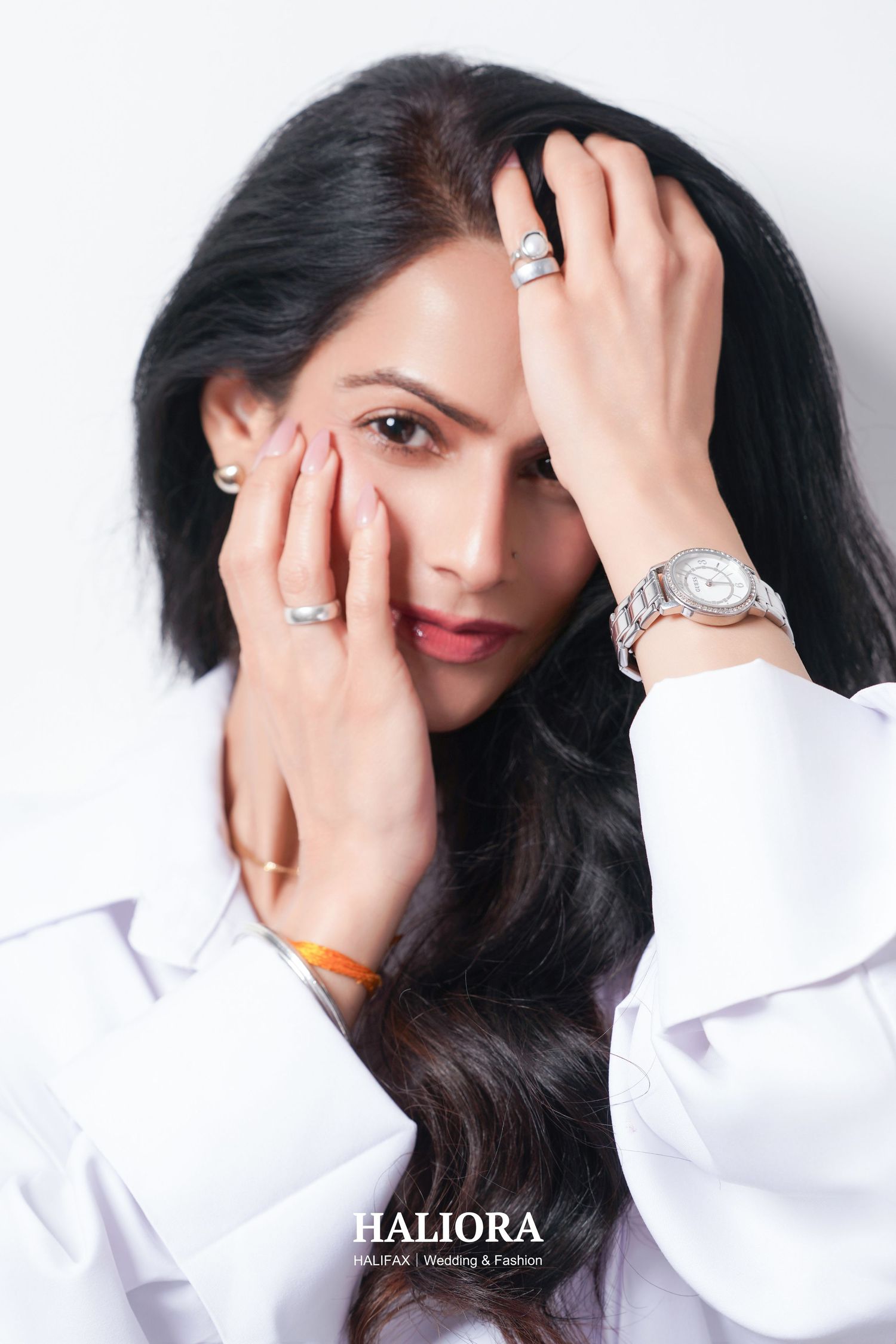 Close-up portrait of woman with dark hair framing face with hands, wearing silver rings and watch, white shirt.