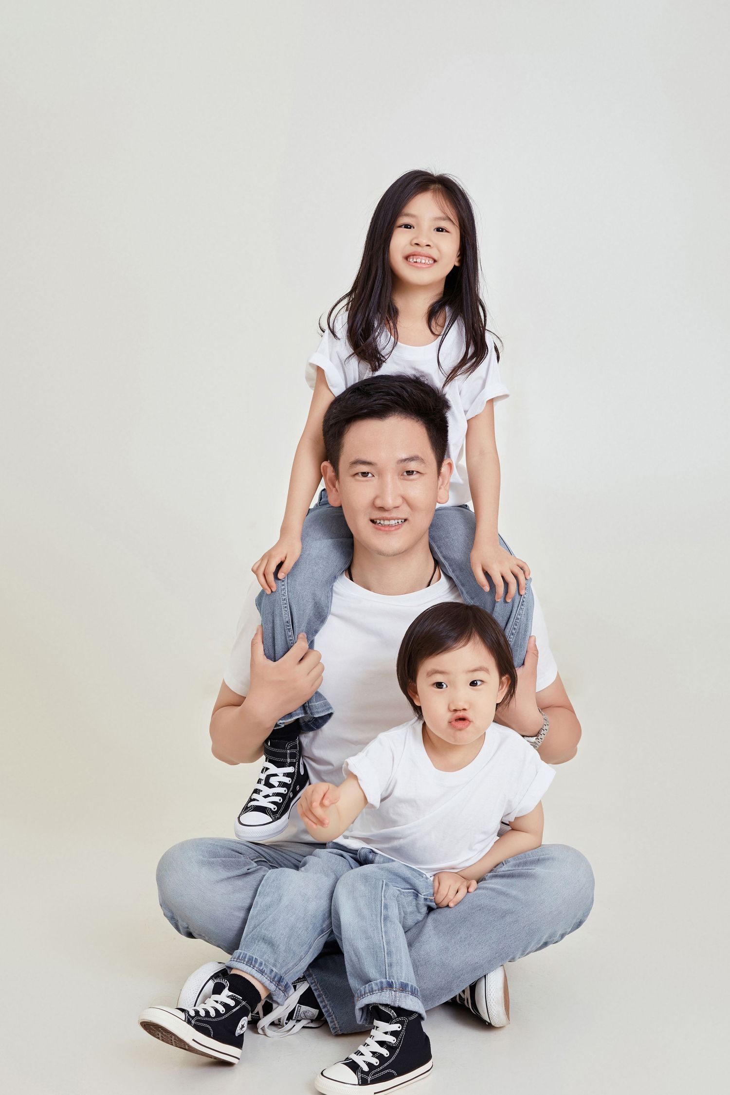 Asian father sitting cross-legged with young daughter on his shoulders and toddler son in matching white tees and jeans.