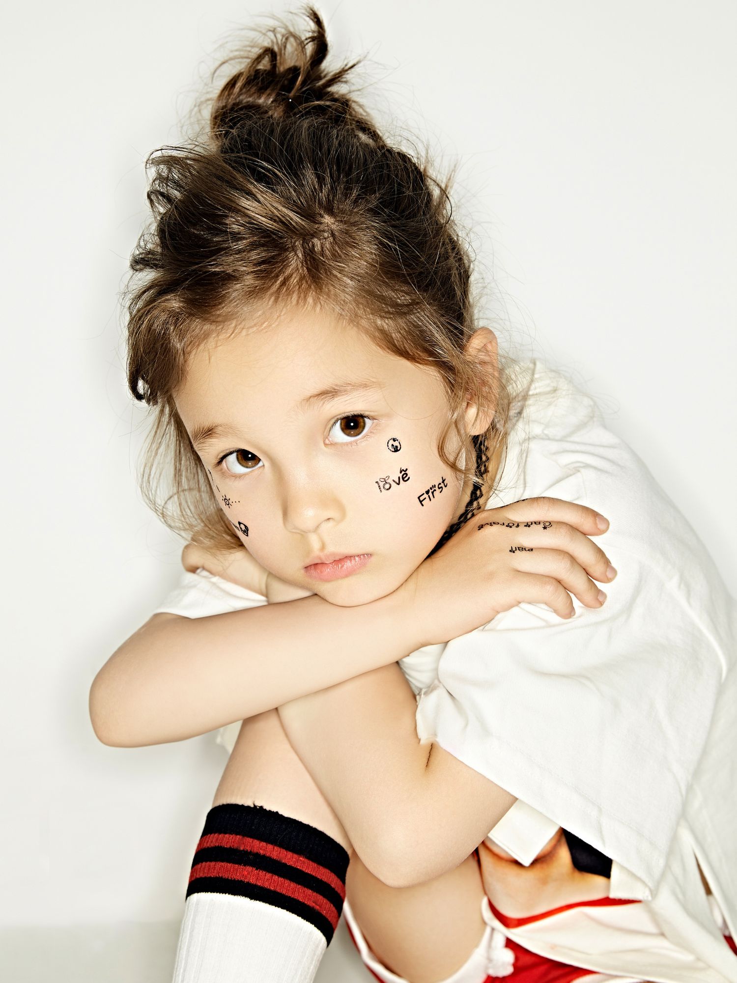 Young child with messy bun hairstyle, face stickers, white shirt, and striped knee-high socks posing thoughtfully.