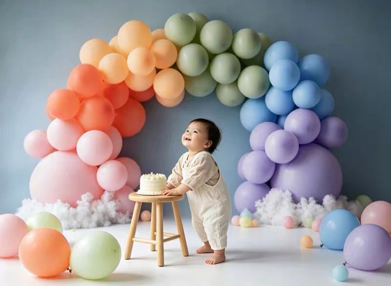 Baby smiling at first birthday cake smash surrounded by pastel rainbow balloon arch and colorful balloons on blue backdrop.