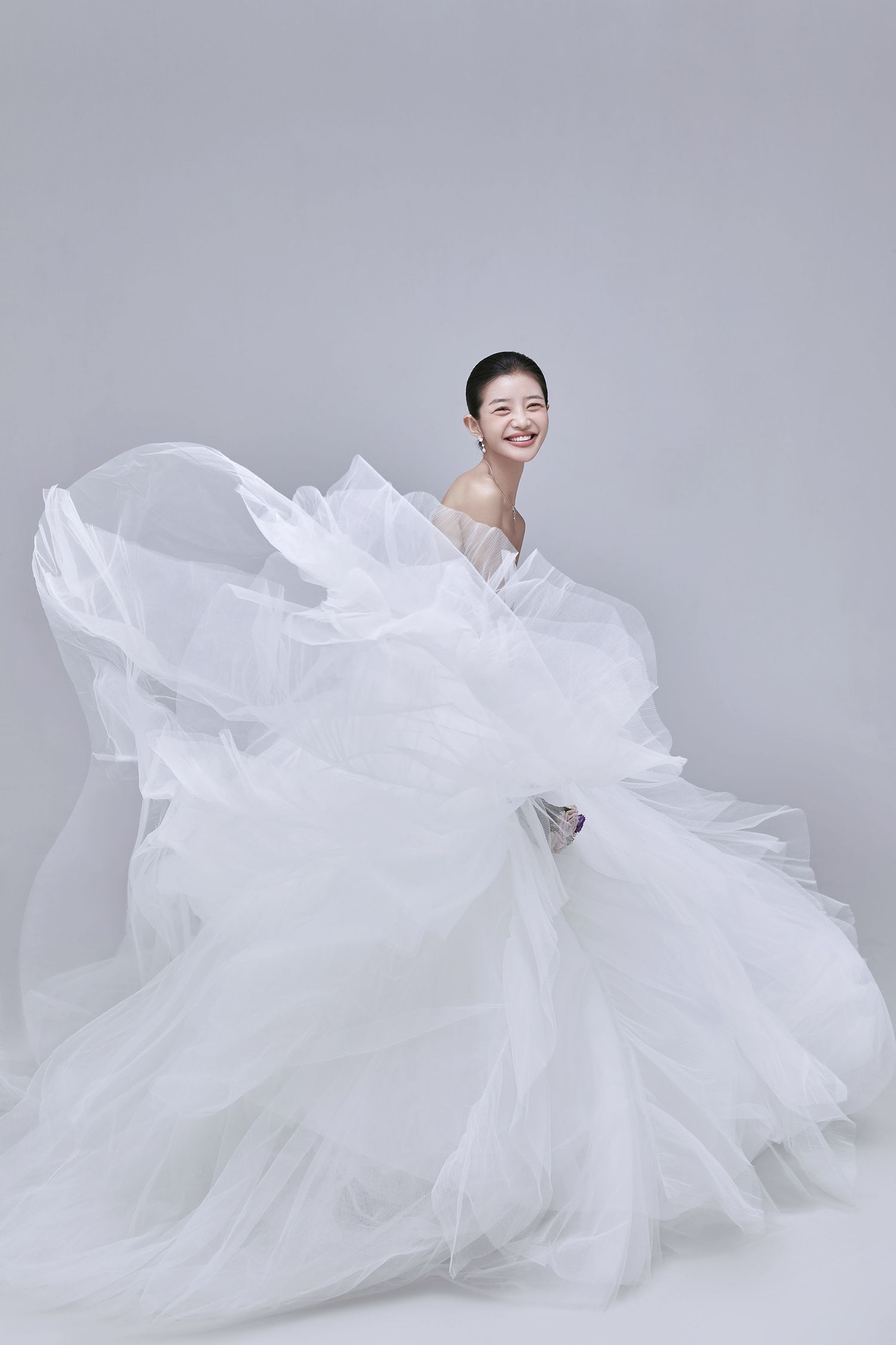 A smiling bride twirls in a flowing white ruffled tulle wedding gown against a gray background.