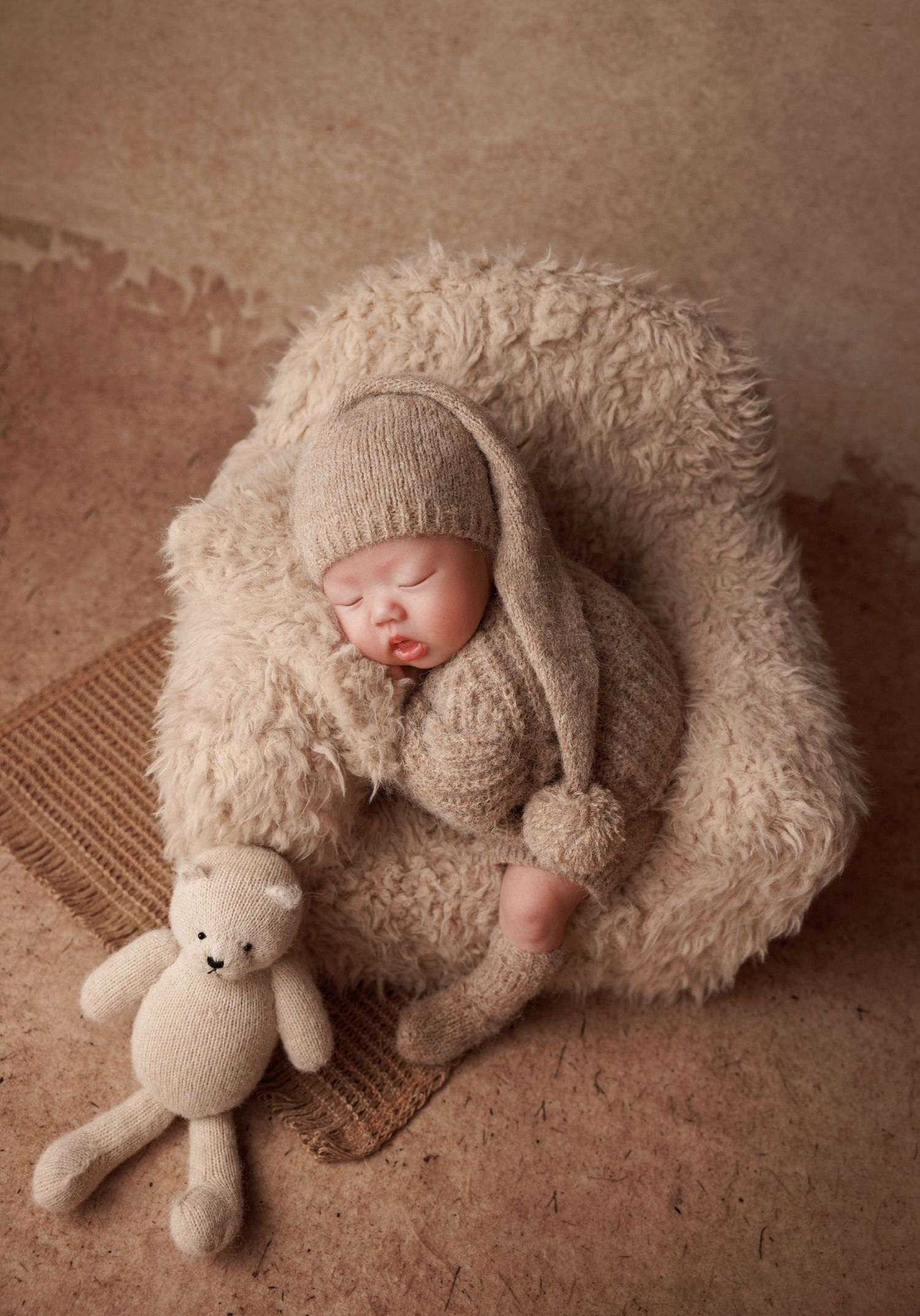 Newborn baby in beige knit outfit and pointed hat sleeping in fluffy cream bowl beside knit teddy bear.