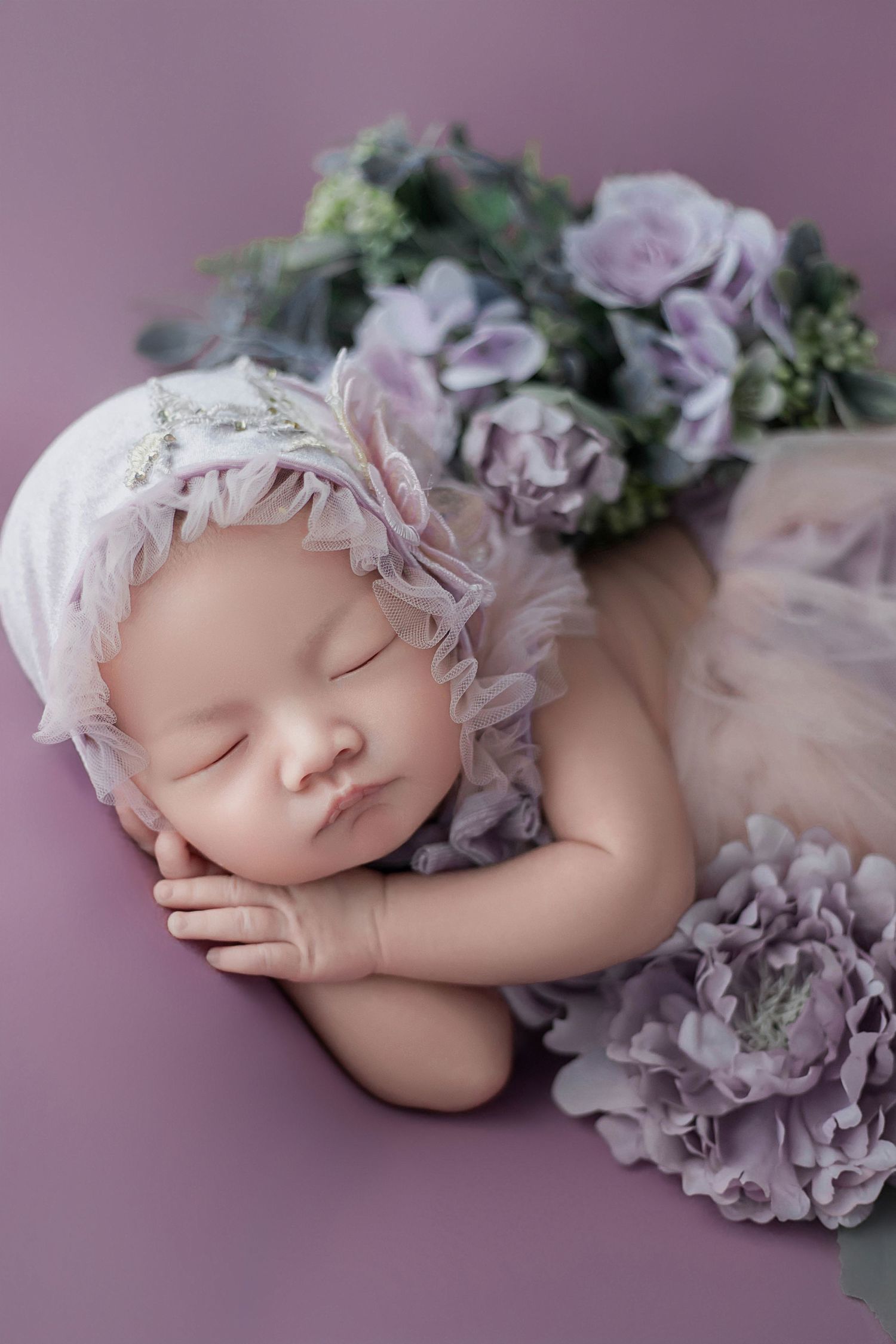 Sleeping newborn baby in white lace bonnet resting on purple background with lavender floral arrangement.