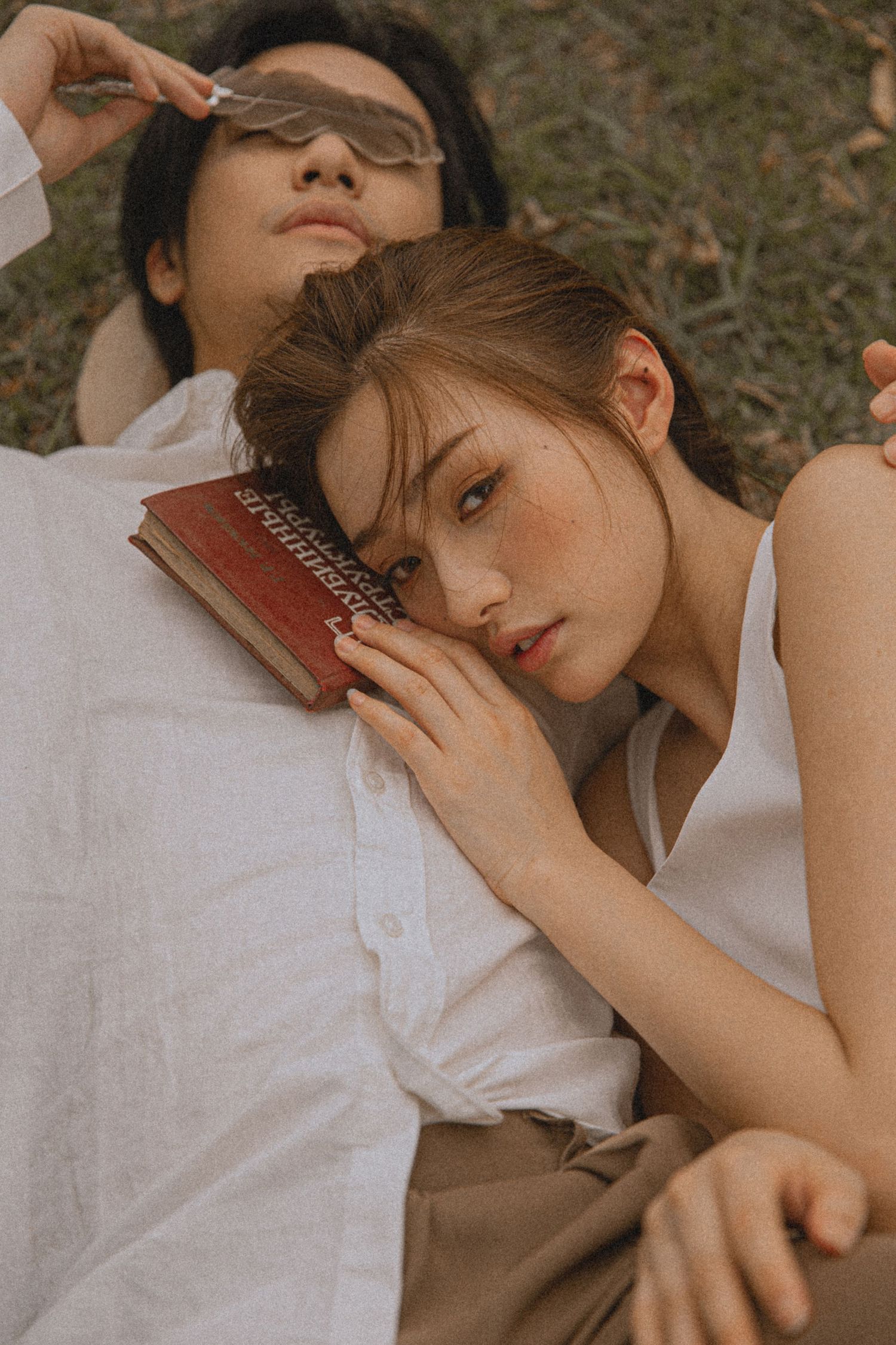 Young couple lying on grass, woman resting head on man's chest holding a red book outdoors.