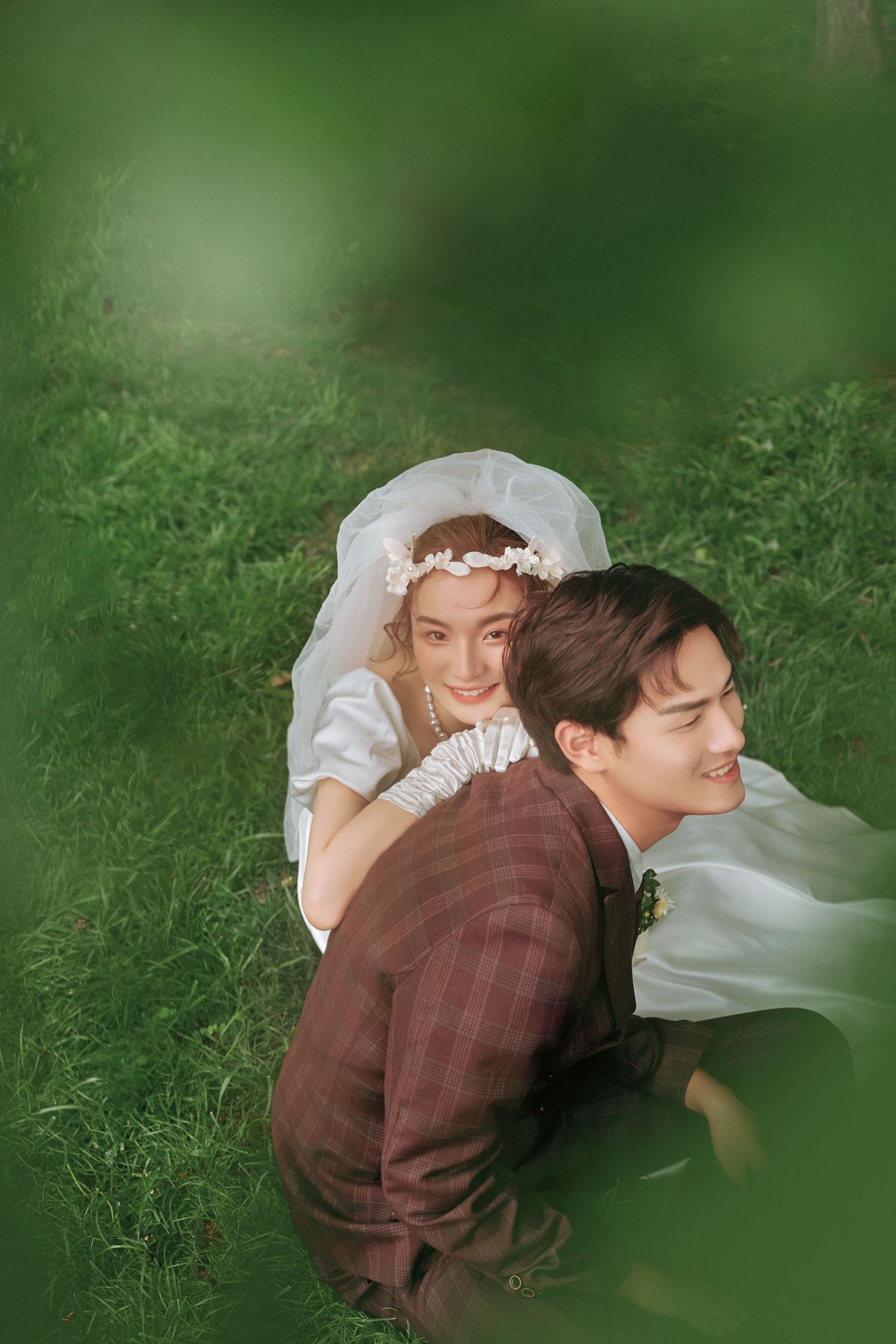 Bride and groom lying on green grass laughing together, bride wearing floral crown and white veil.