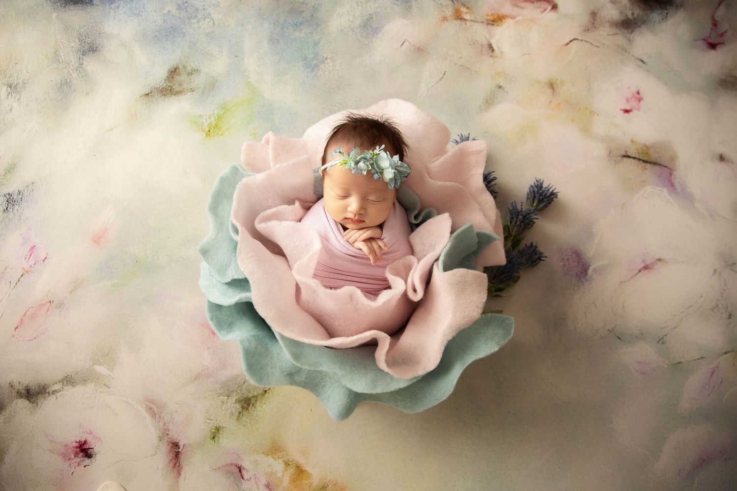 Newborn baby sleeping in a pink wrap with floral headband, posed in a teal flower bowl on pastel backdrop.