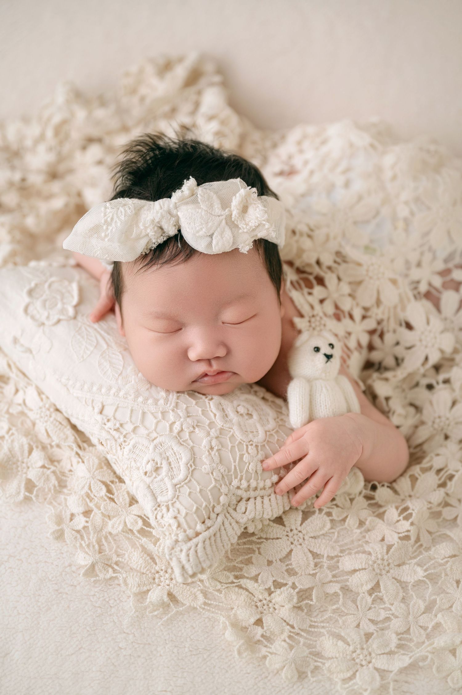 Sleeping newborn baby girl with floral headband cuddling small white knit bear.