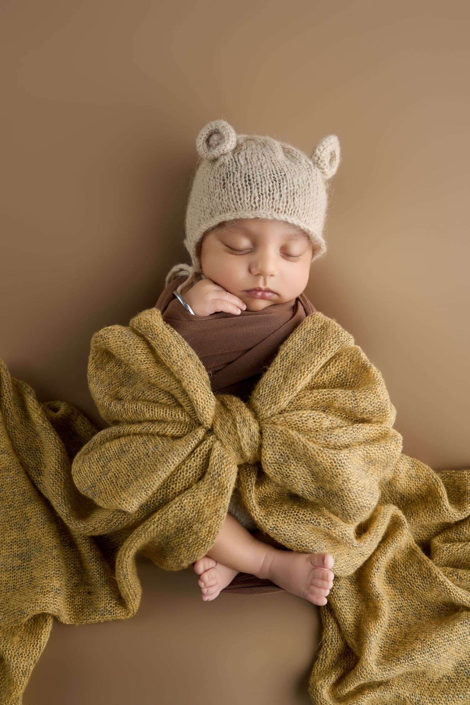 Sleeping newborn baby wrapped in golden knit blanket wearing a cream bear ear hat on beige background.