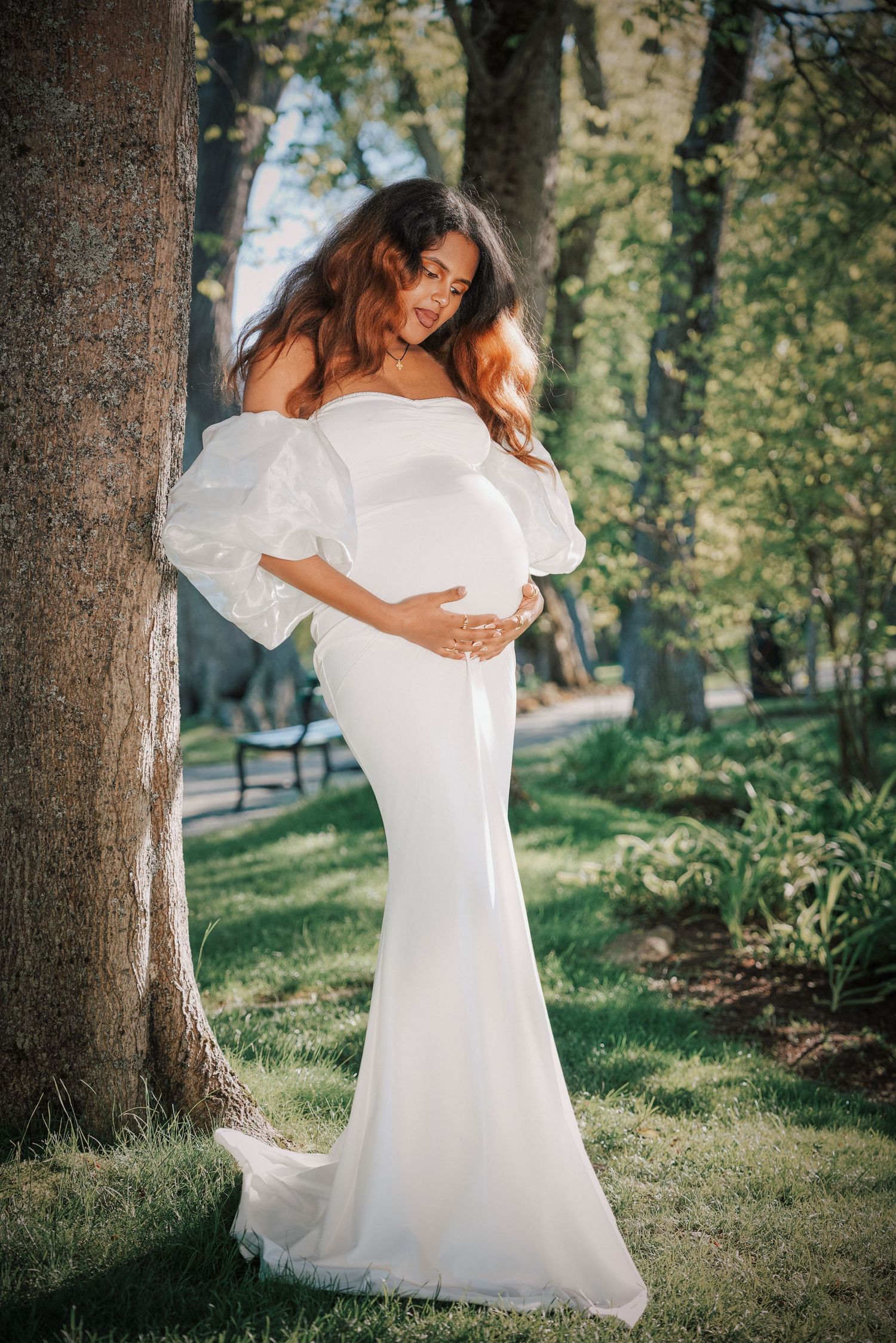 Pregnant woman in white off-shoulder gown with puff sleeves poses by tree in lush green park.