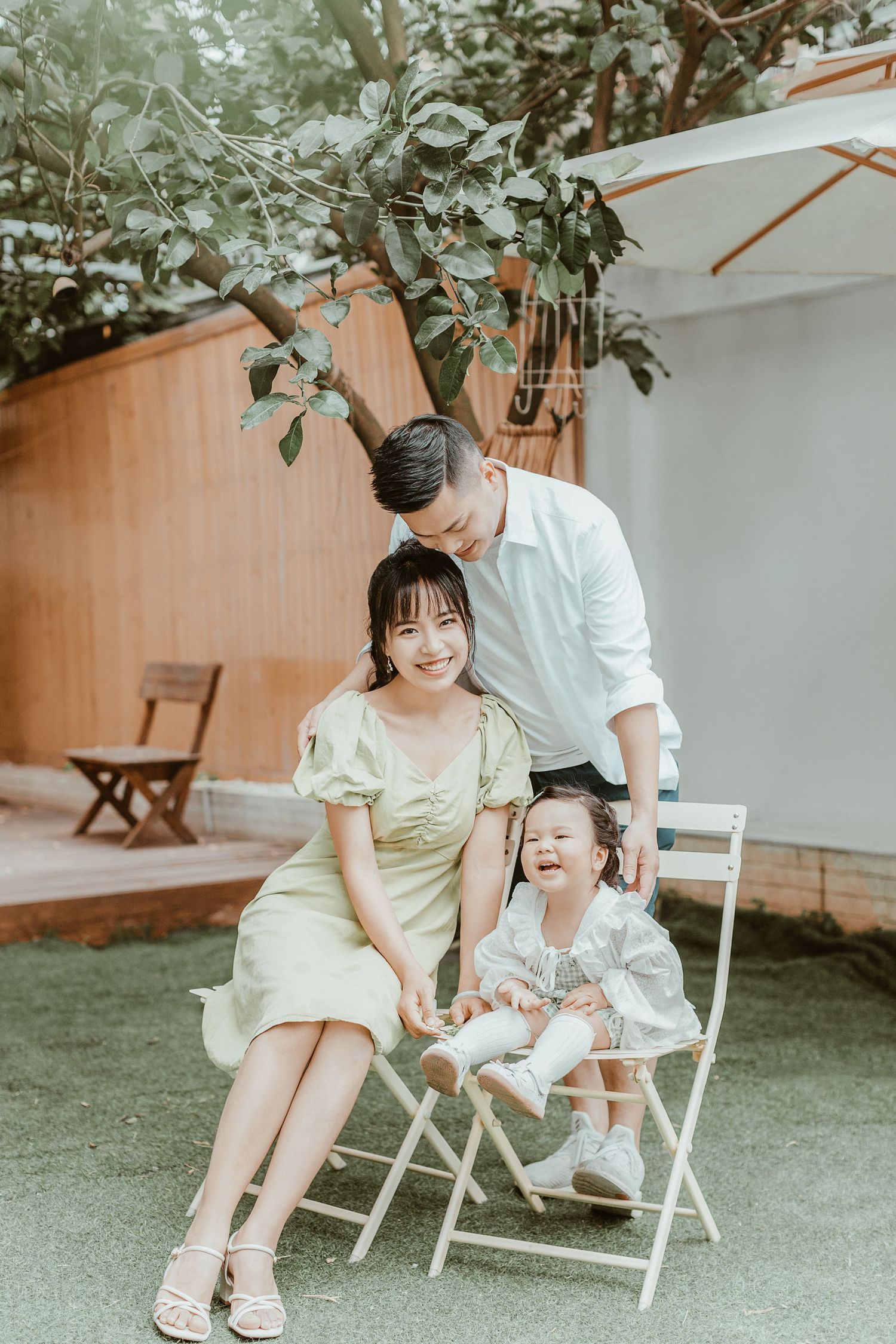 Happy Asian family with young toddler daughter poses outdoors in a lush garden setting.