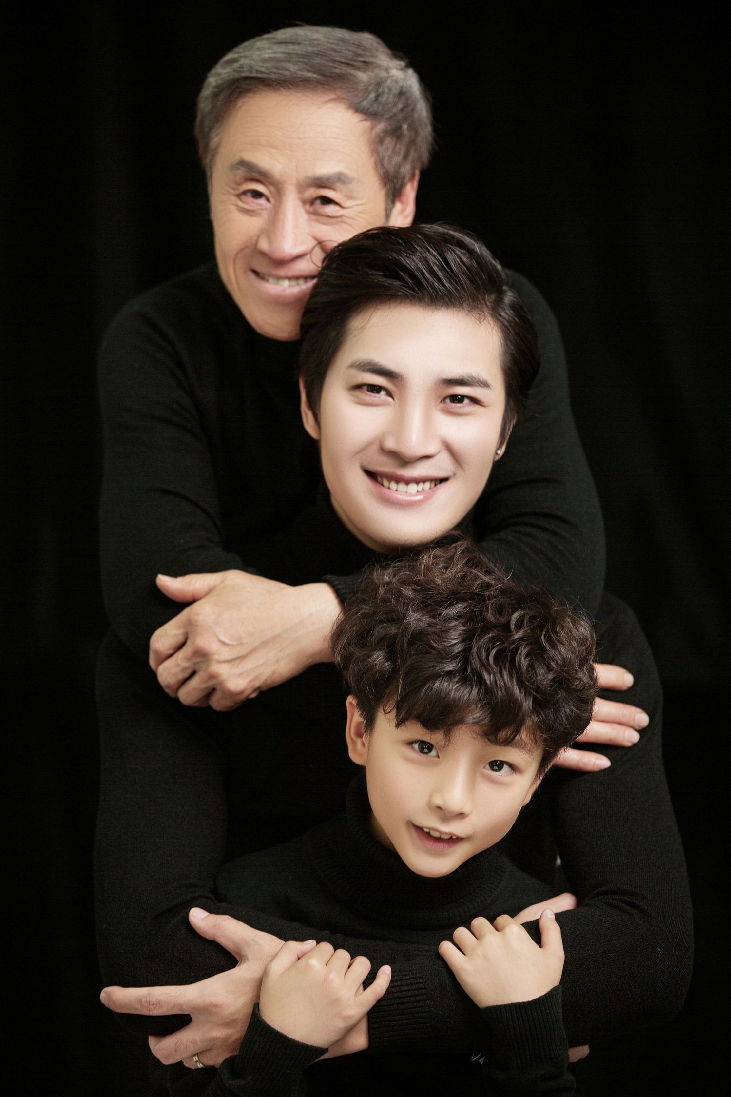 Three generations of Asian males pose warmly together in matching black turtlenecks in a studio portrait.