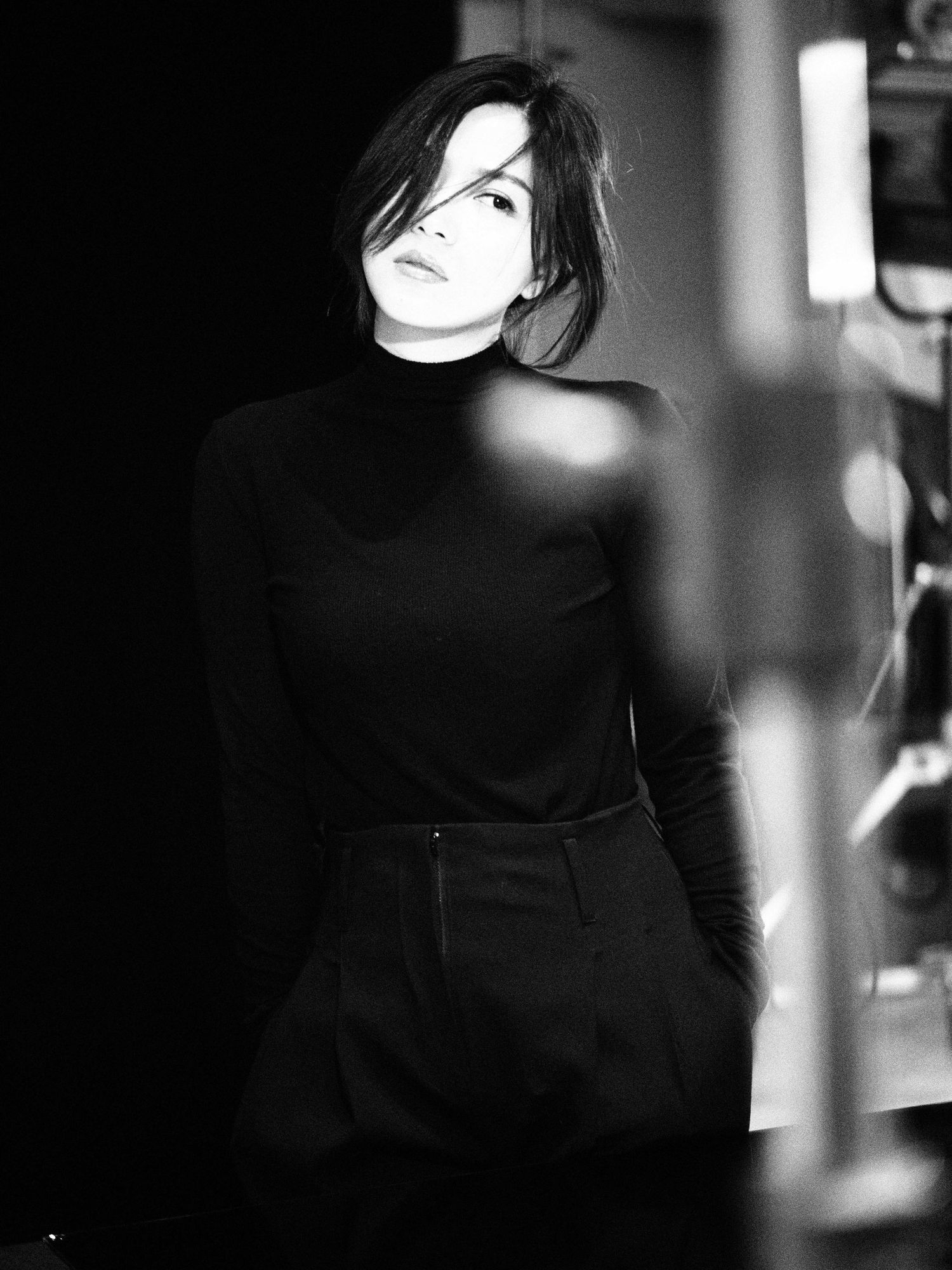 Black and white fashion portrait of a woman in black turtleneck and skirt posing in dramatic studio lighting.