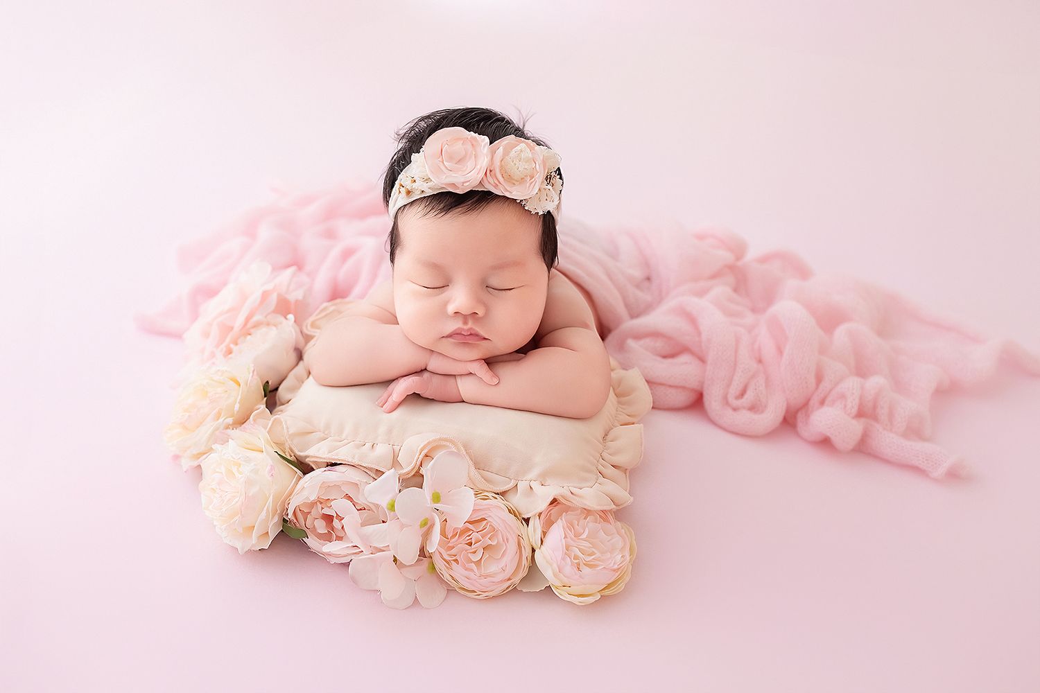 Peaceful newborn baby girl wearing floral headband sleeping on pillow in wooden box with pink roses.