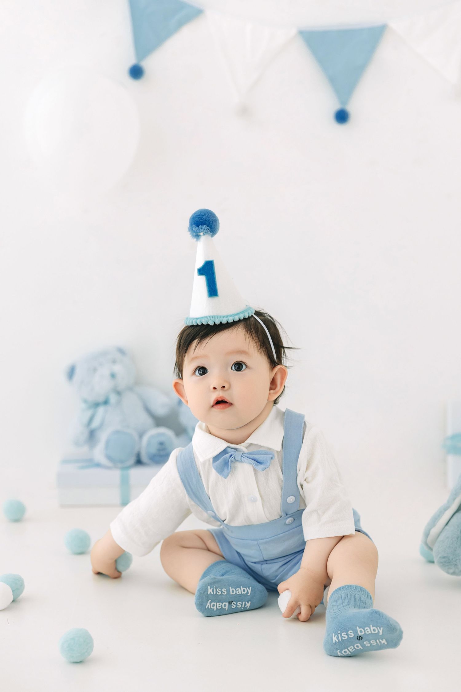 Adorable baby boy in blue suspenders and party hat celebrating his 1st birthday in a white decorated studio.