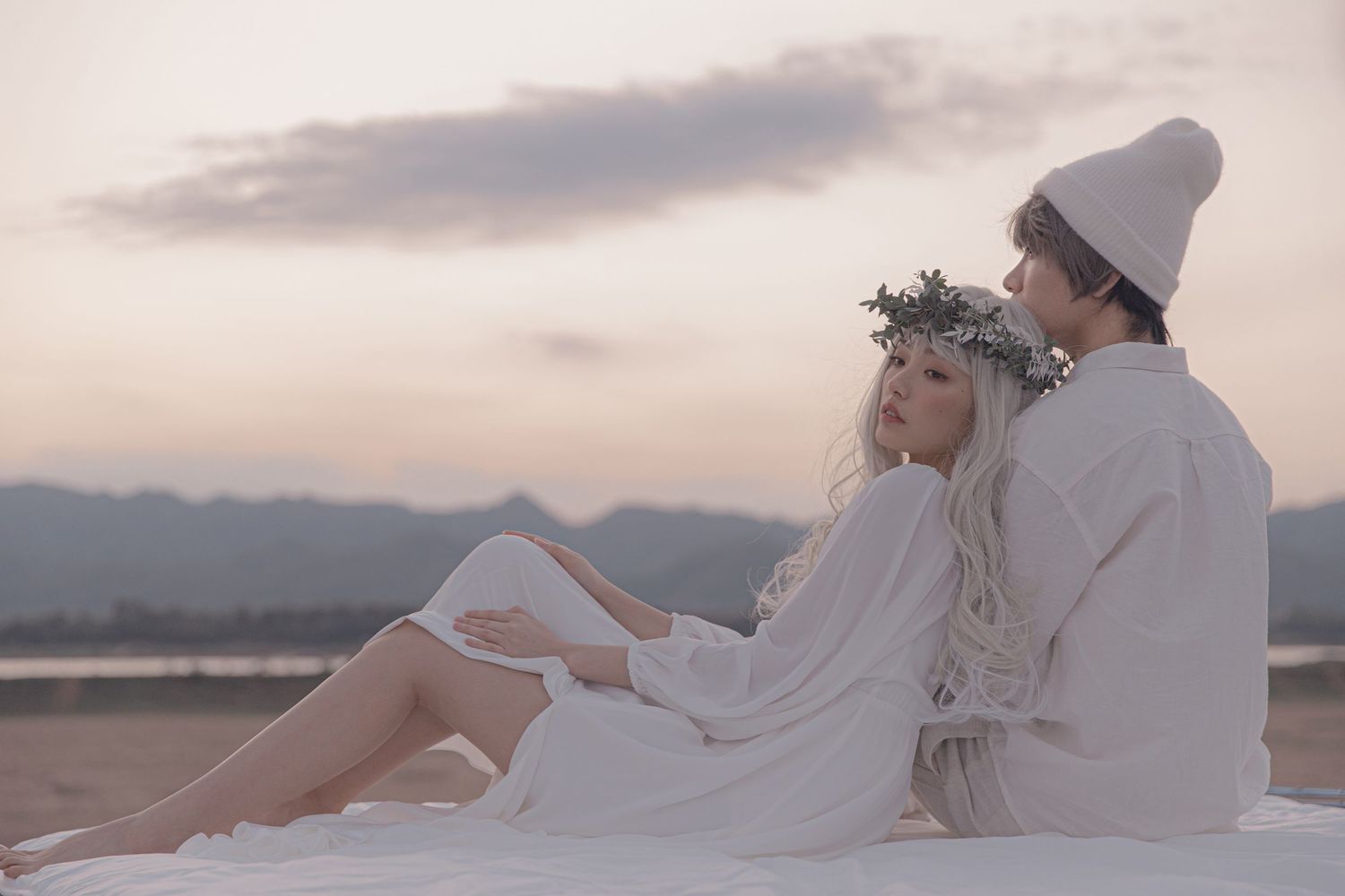 Couple in white outfits sit together at sunset, woman in floral crown and man in white beanie.