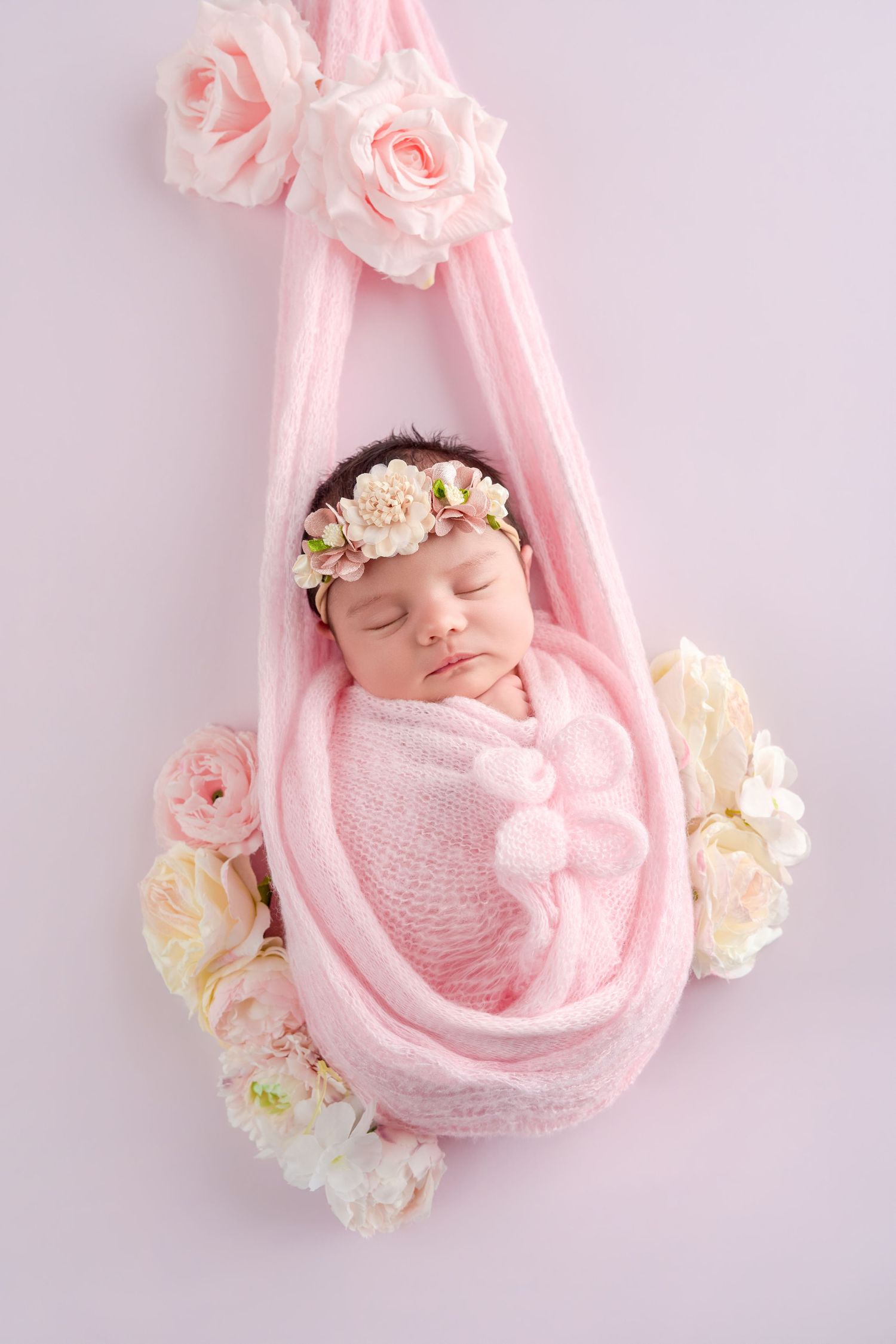 Sleeping newborn baby wrapped in pink knit blanket with floral headband, surrounded by roses in a hanging cradle.