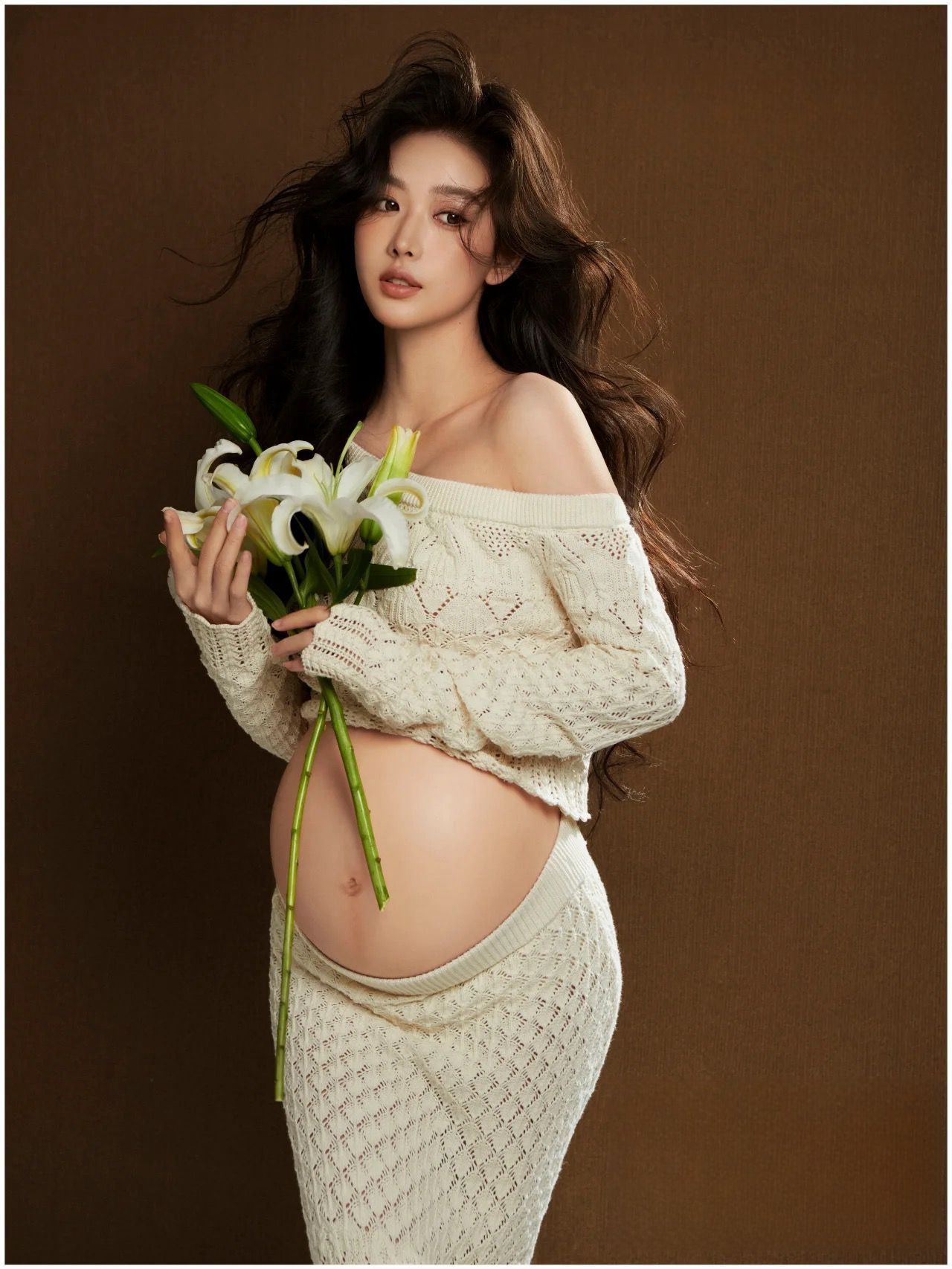 Glowing pregnant model in cream knit maternity set holding white lily bouquet against brown backdrop.