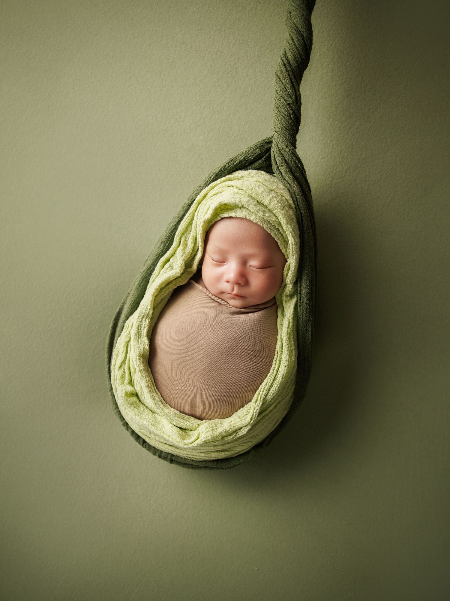 Newborn baby swaddled in green wrap sleeping inside hanging avocado-shaped pod on green background.