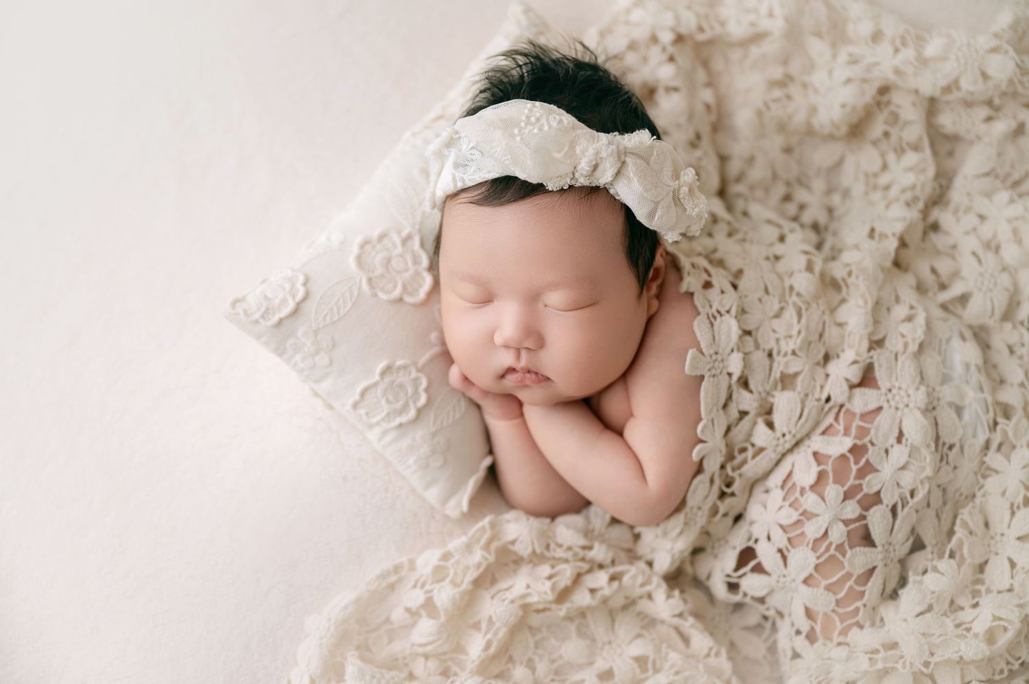 Newborn baby with dark hair sleeping on lace pillow wrapped in cream floral lace blanket with bow headband.