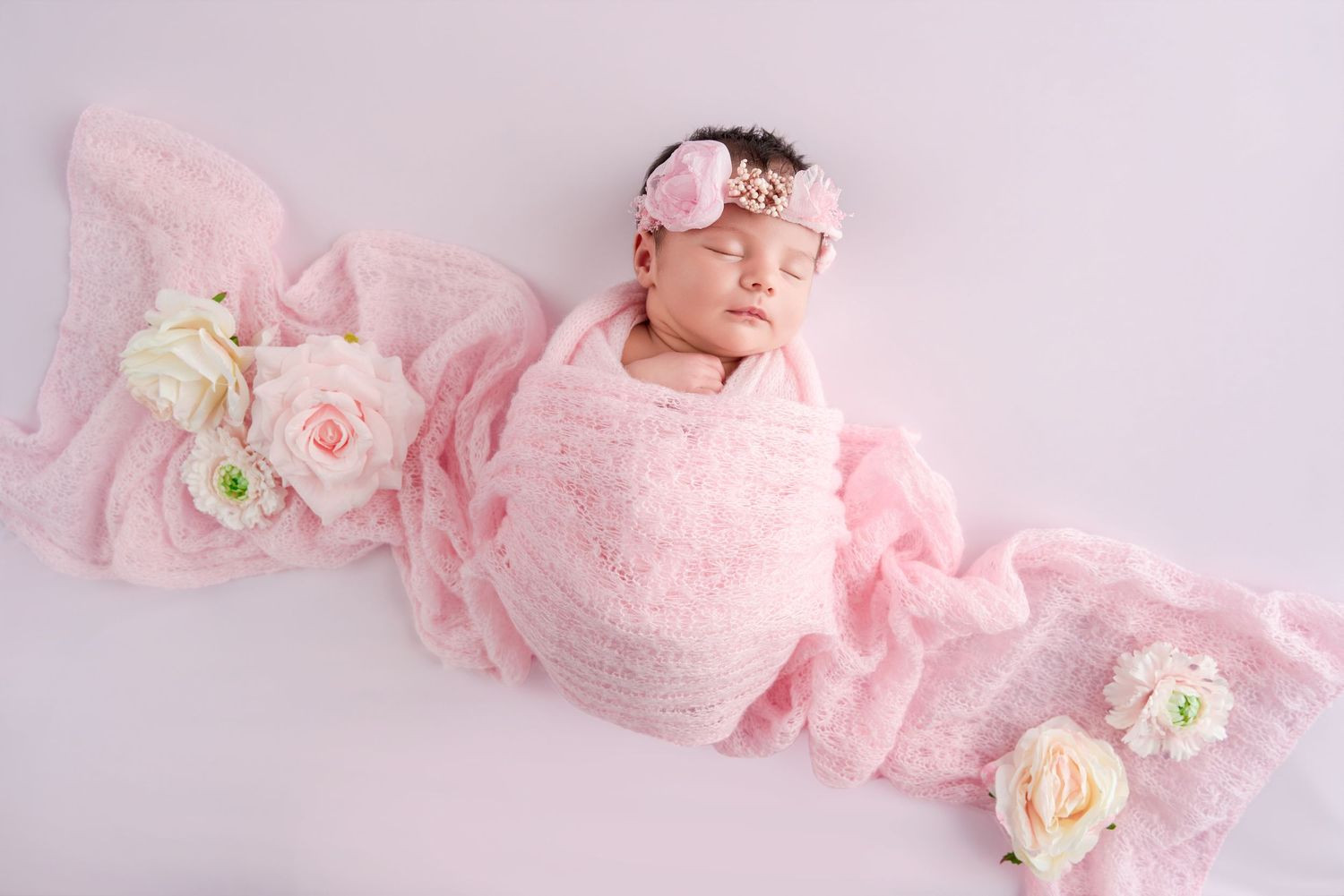 Newborn baby girl wrapped in pink swaddling cloth with floral headband, surrounded by roses on white background.