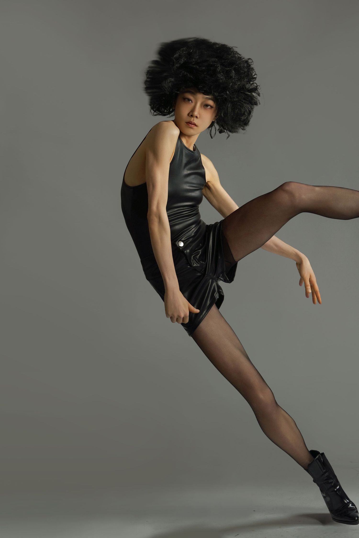 Asian female model in black leather mini dress and afro wig strikes a dynamic dance pose on gray background.