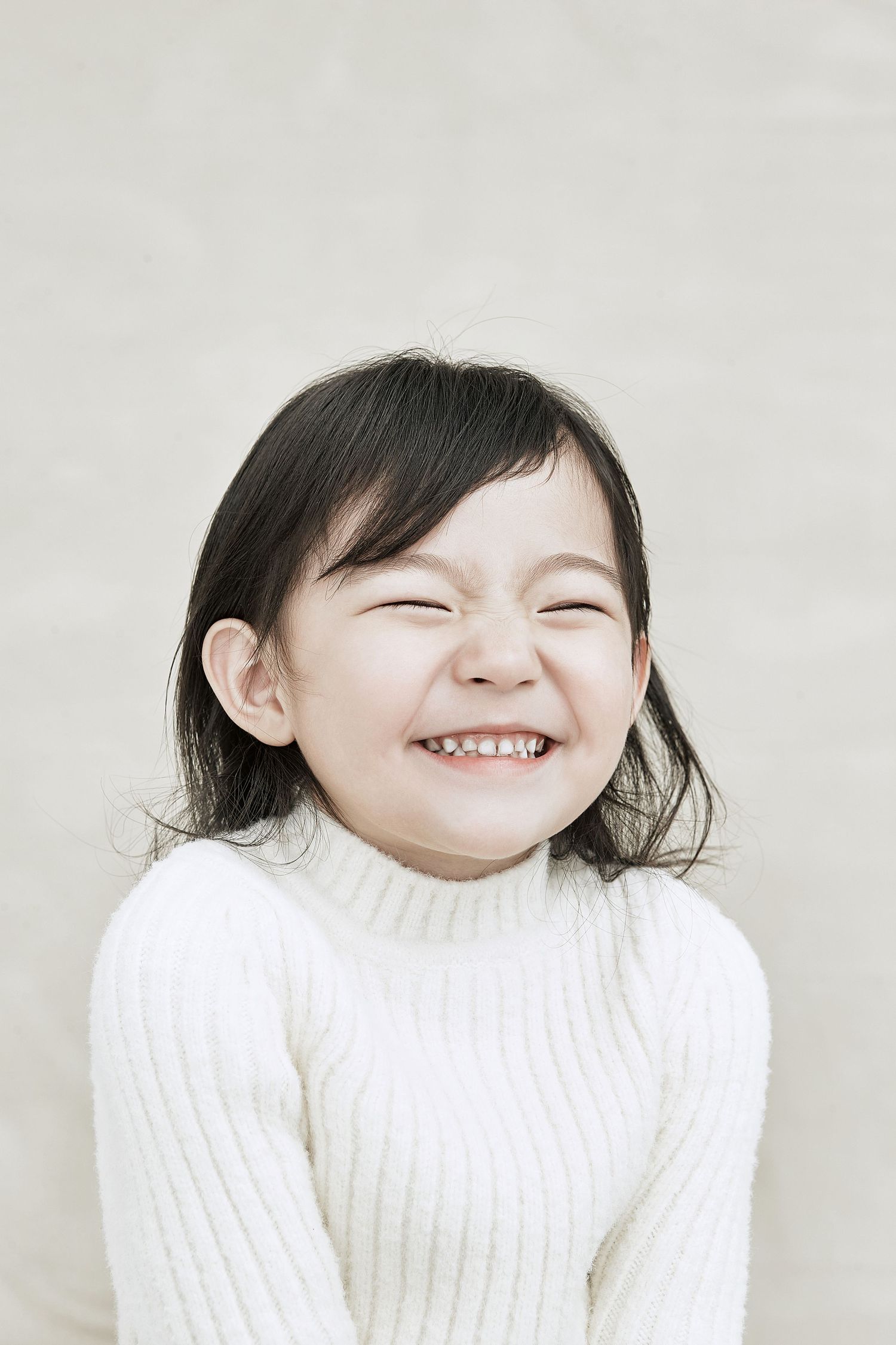 A young Asian girl in a white knit sweater laughing with eyes closed against a beige background.