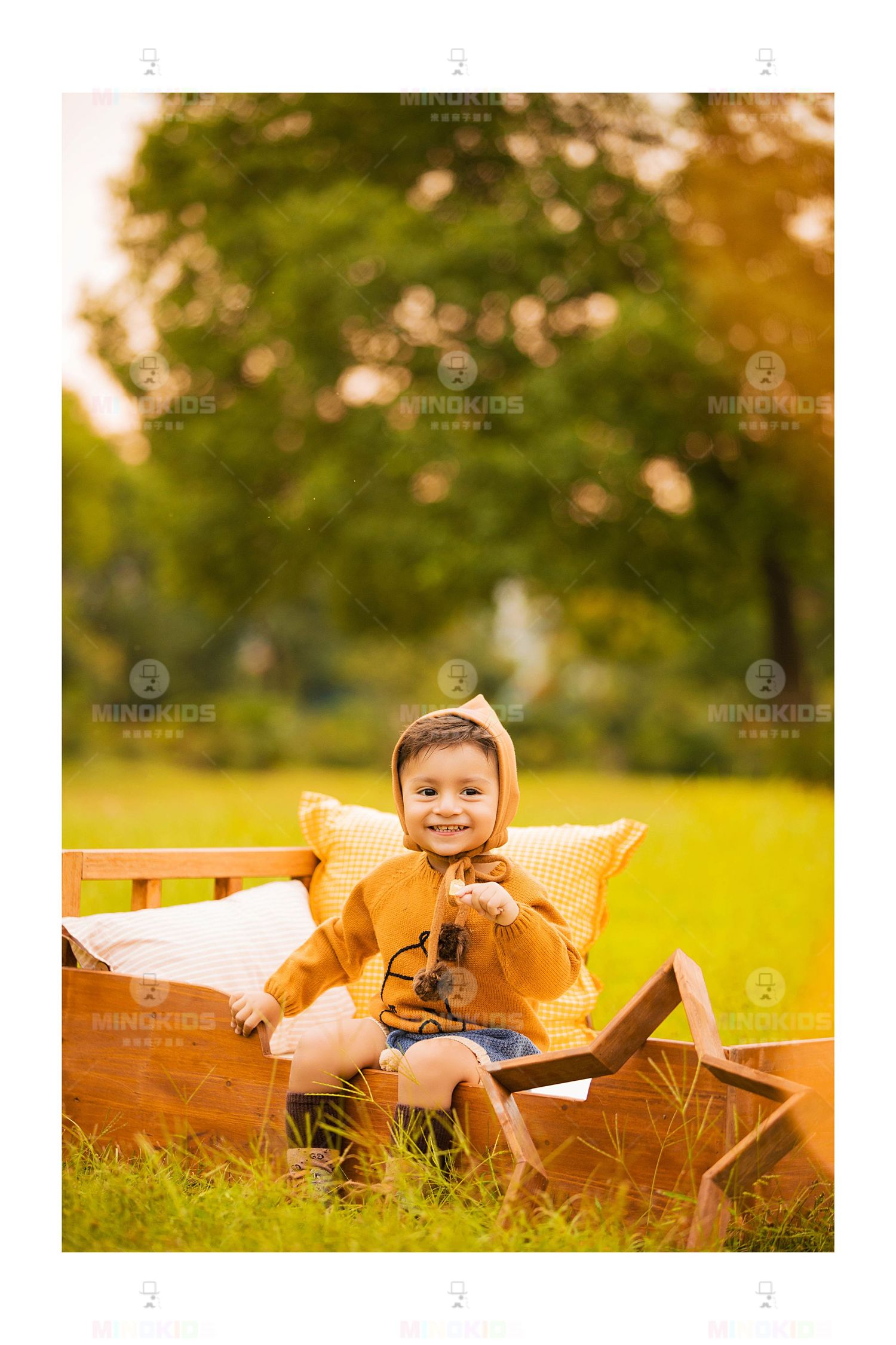 Smiling toddler in orange jacket holds toy binoculars on wooden bed prop in autumn park setting.