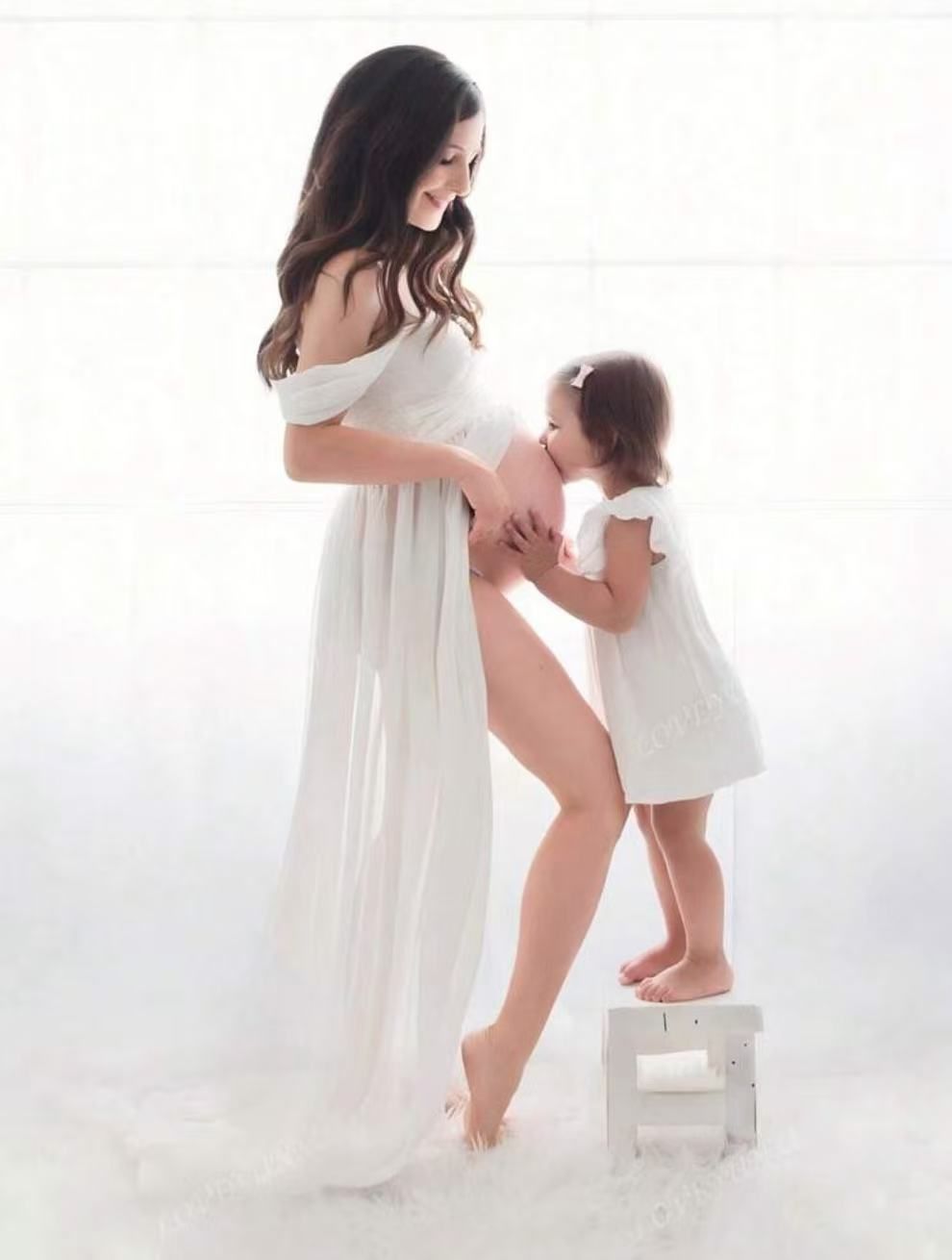 Pregnant woman in white off-shoulder chiffon maternity gown smiling at toddler daughter in white studio.