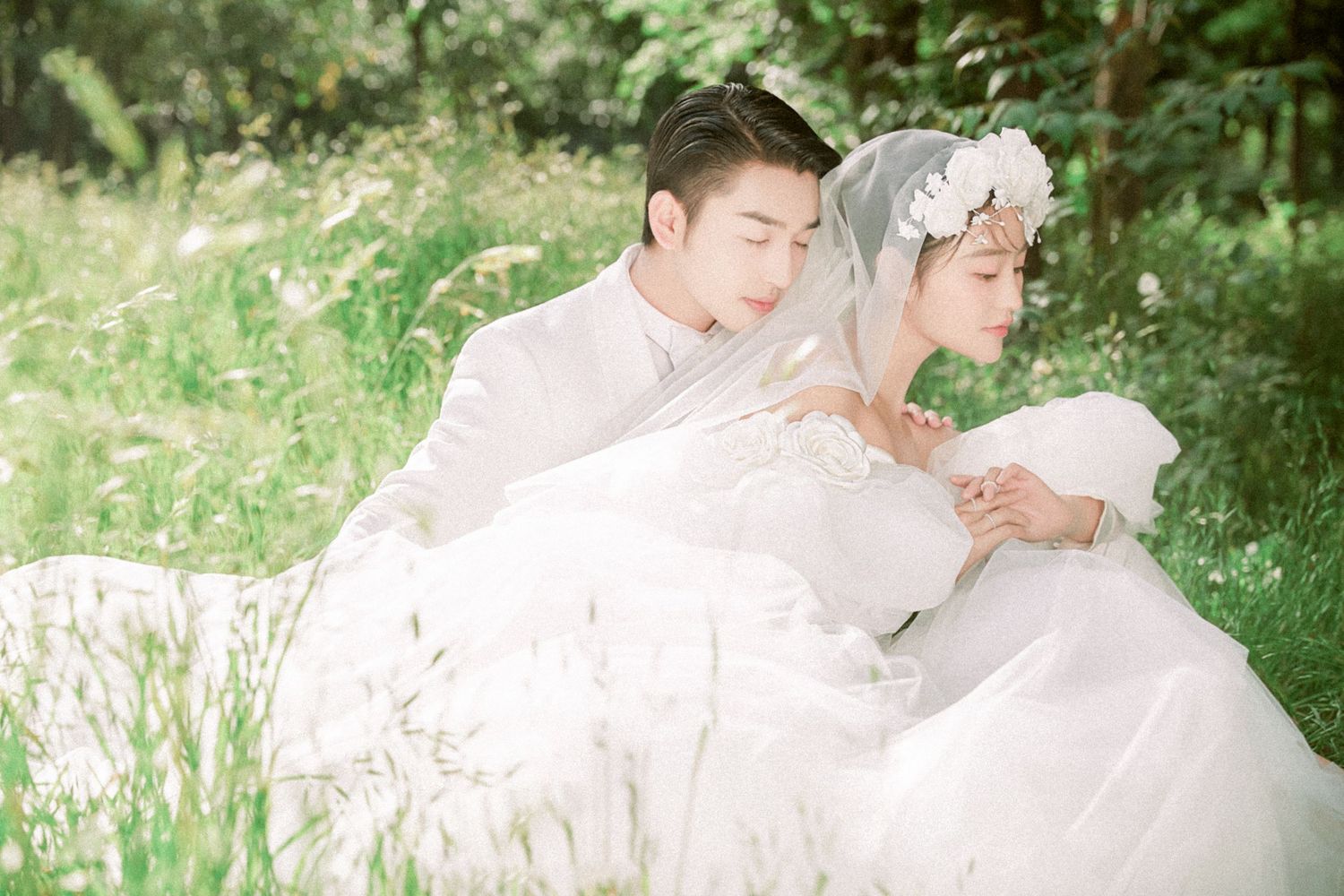 Couple in white wedding attire sit back-to-back in a lush meadow with wildflowers.