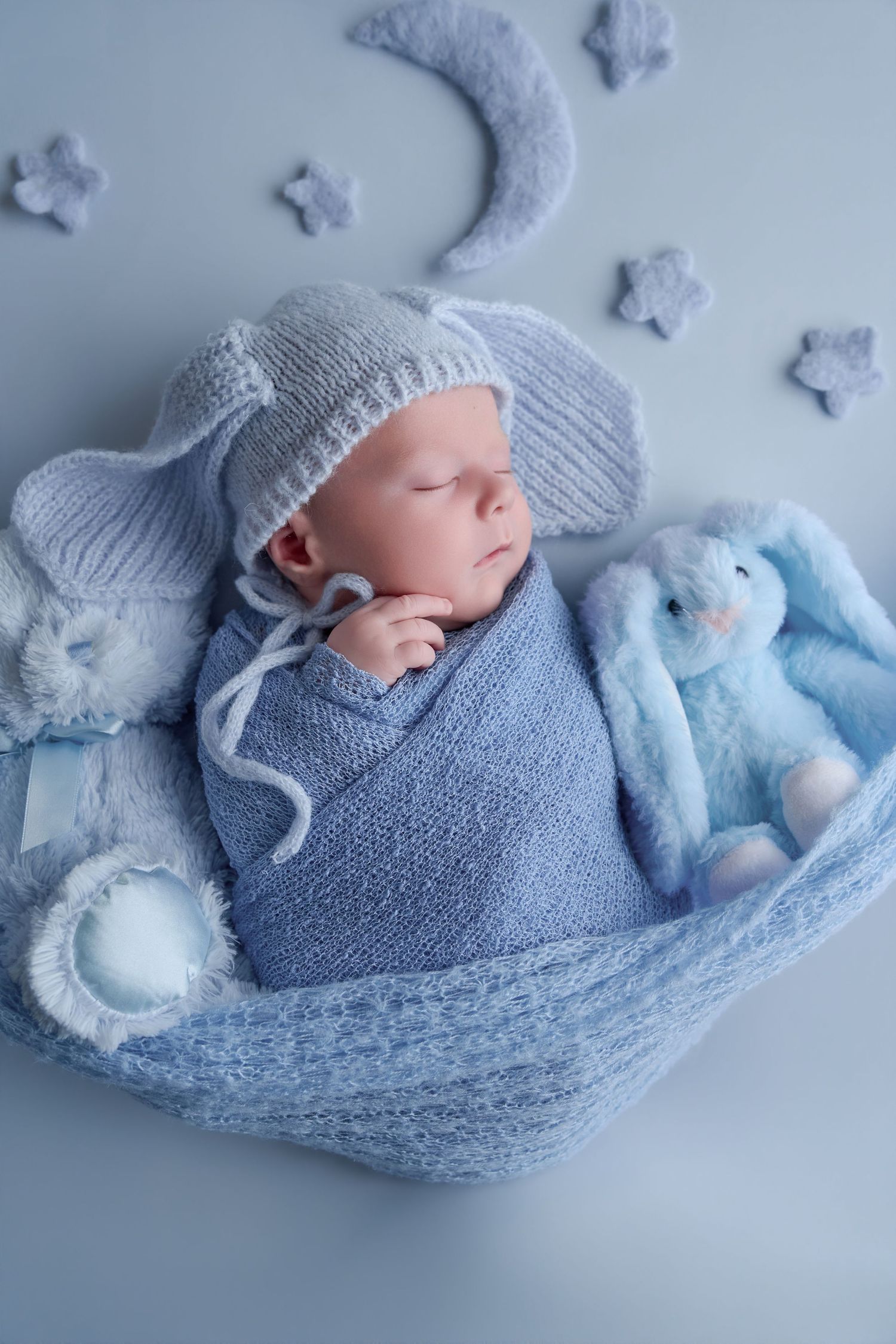 Sleeping newborn baby wrapped in blue knit blanket with bunny toy, moon and stars, wearing a cozy knitted hat.