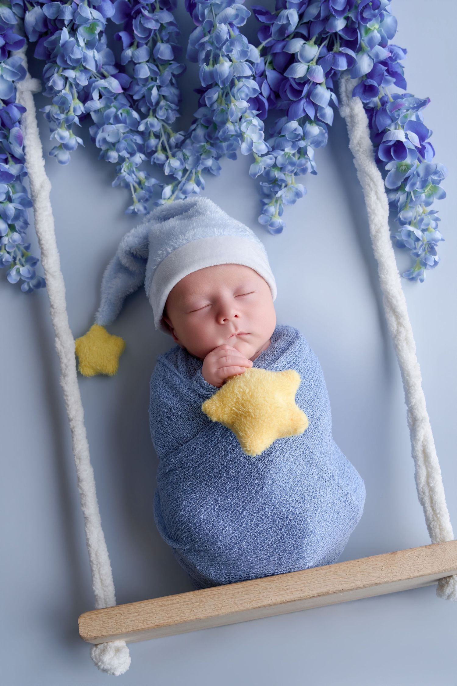 Newborn baby swaddled in blue wrap wearing sleep hat, holding yellow star, resting on swing with purple flowers.